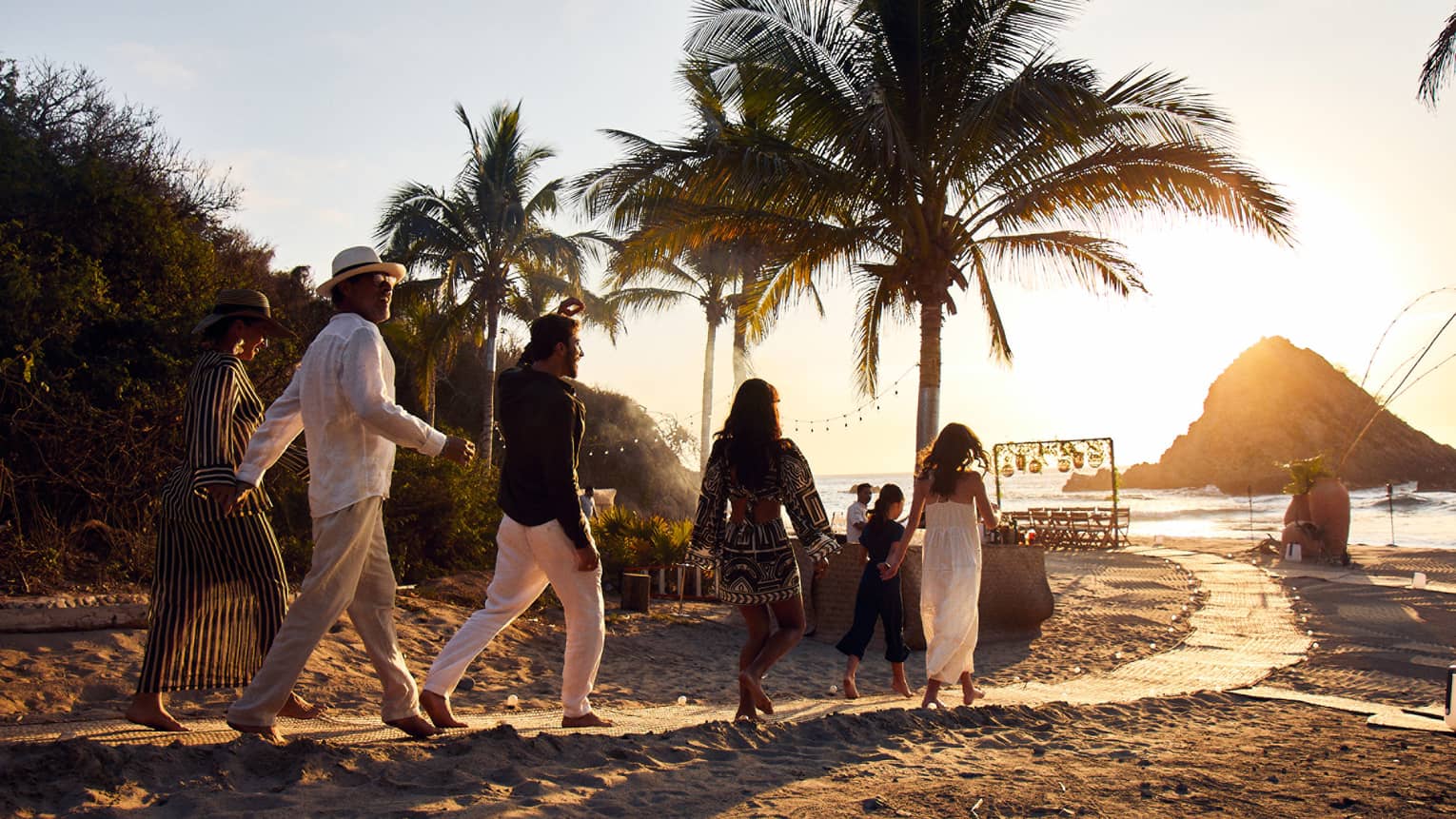 A group of guests walk along a groomed beach path toward a table set beside the water as the sun sets behind a rock outcrop.