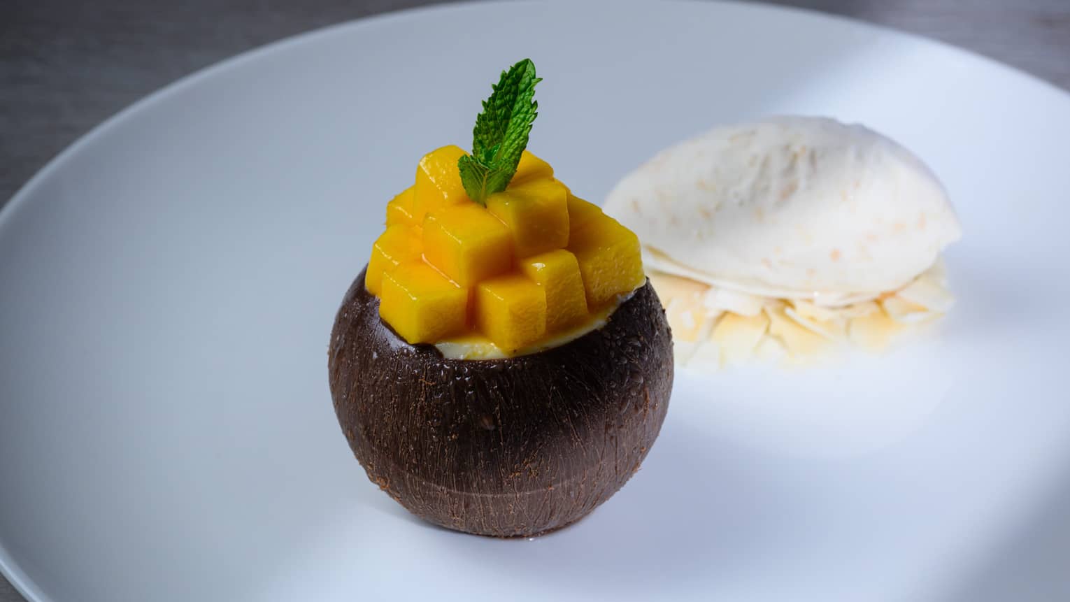 Whole coconut filled with mango cubes on white plate with scoop of vanilla ice cream