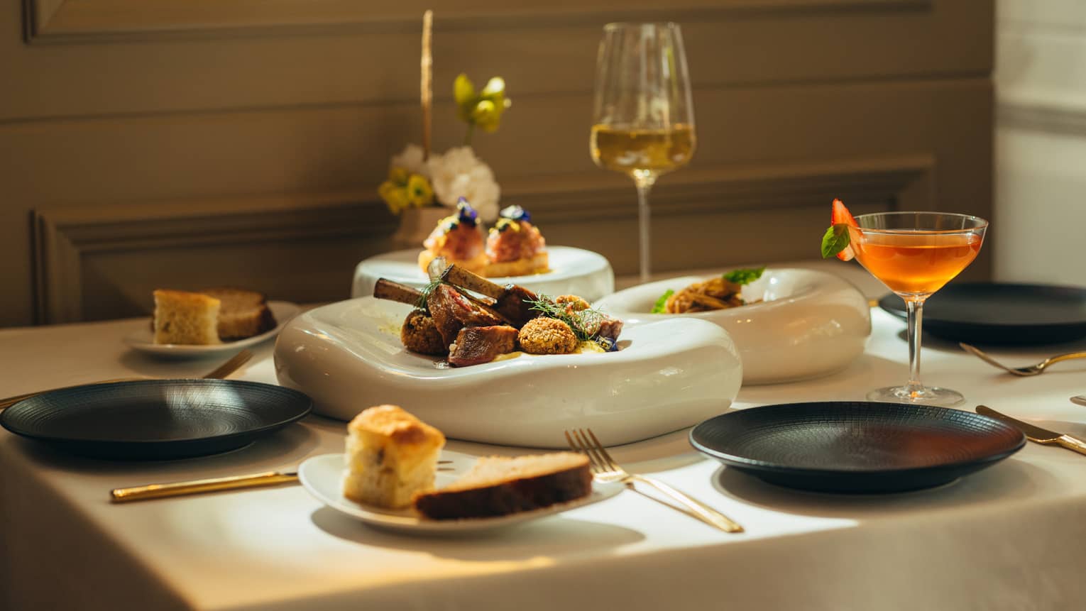 Three white serving platters with food sit in the middle of a white-cloth dining table with three black plates, a glass of white wine, a cocktail in a coupe and two small plates of bread surrounding them