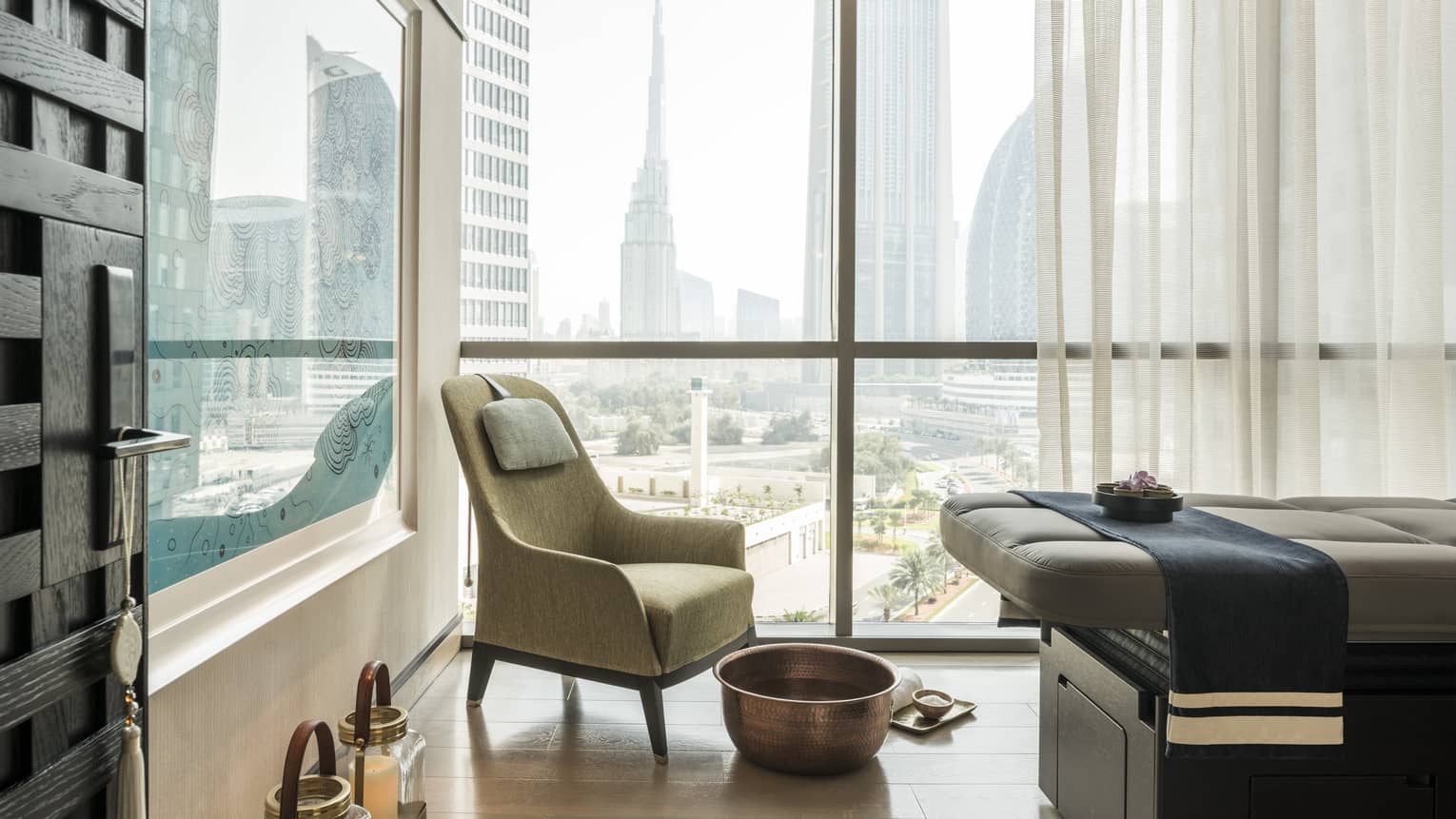Armchair in hotel room corner by sunny window, large brass bowl foot bath, rolled white towel on floor