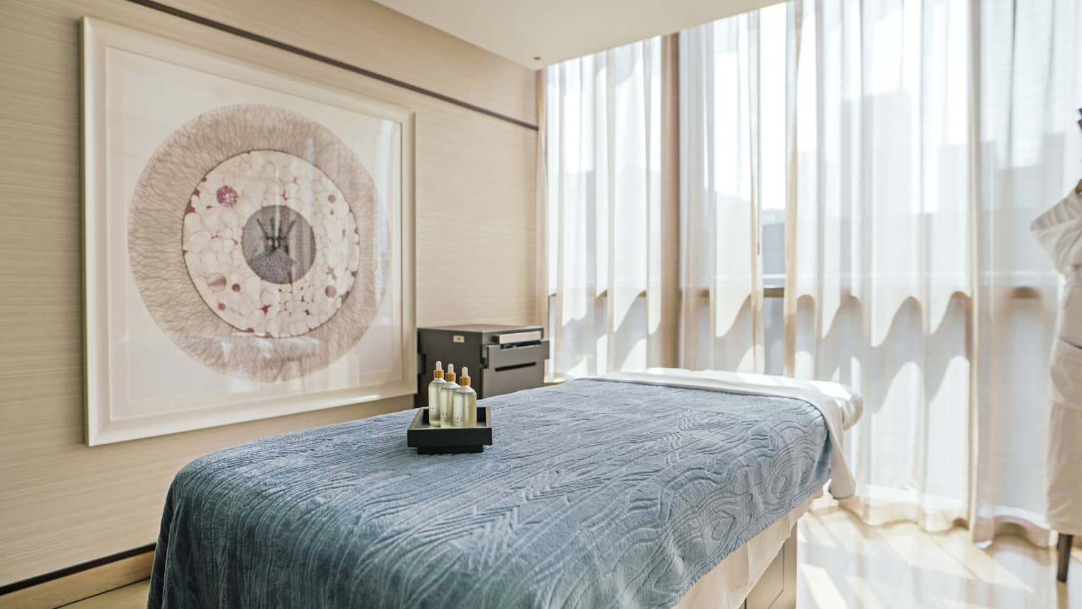 Spa treatment room with white walls, natural light and a massage table topped with a light-blue blanket and a tray of oils