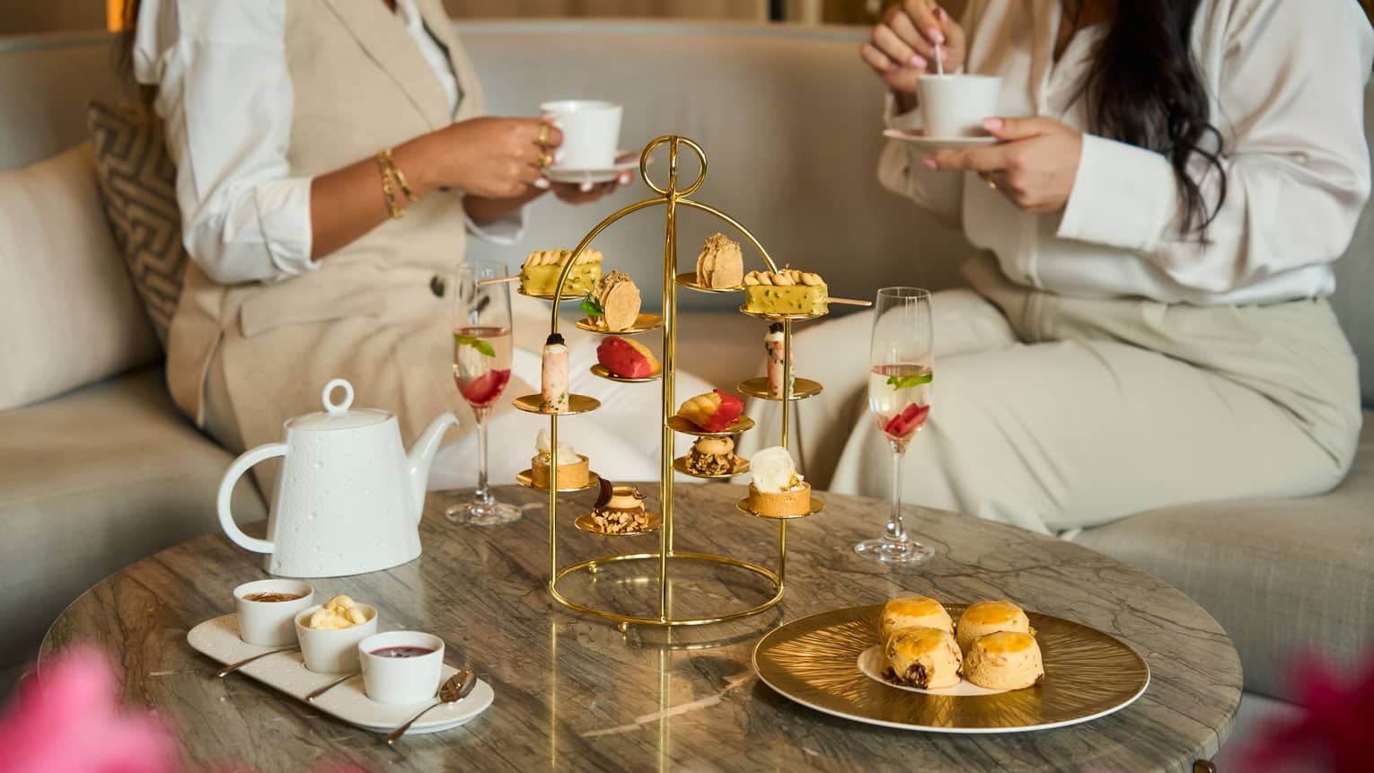 Two people sit on a beige sofa next to a marble coffee table set with a golden tray holding a variety of pastries, a white tea pot and othe small plates of food