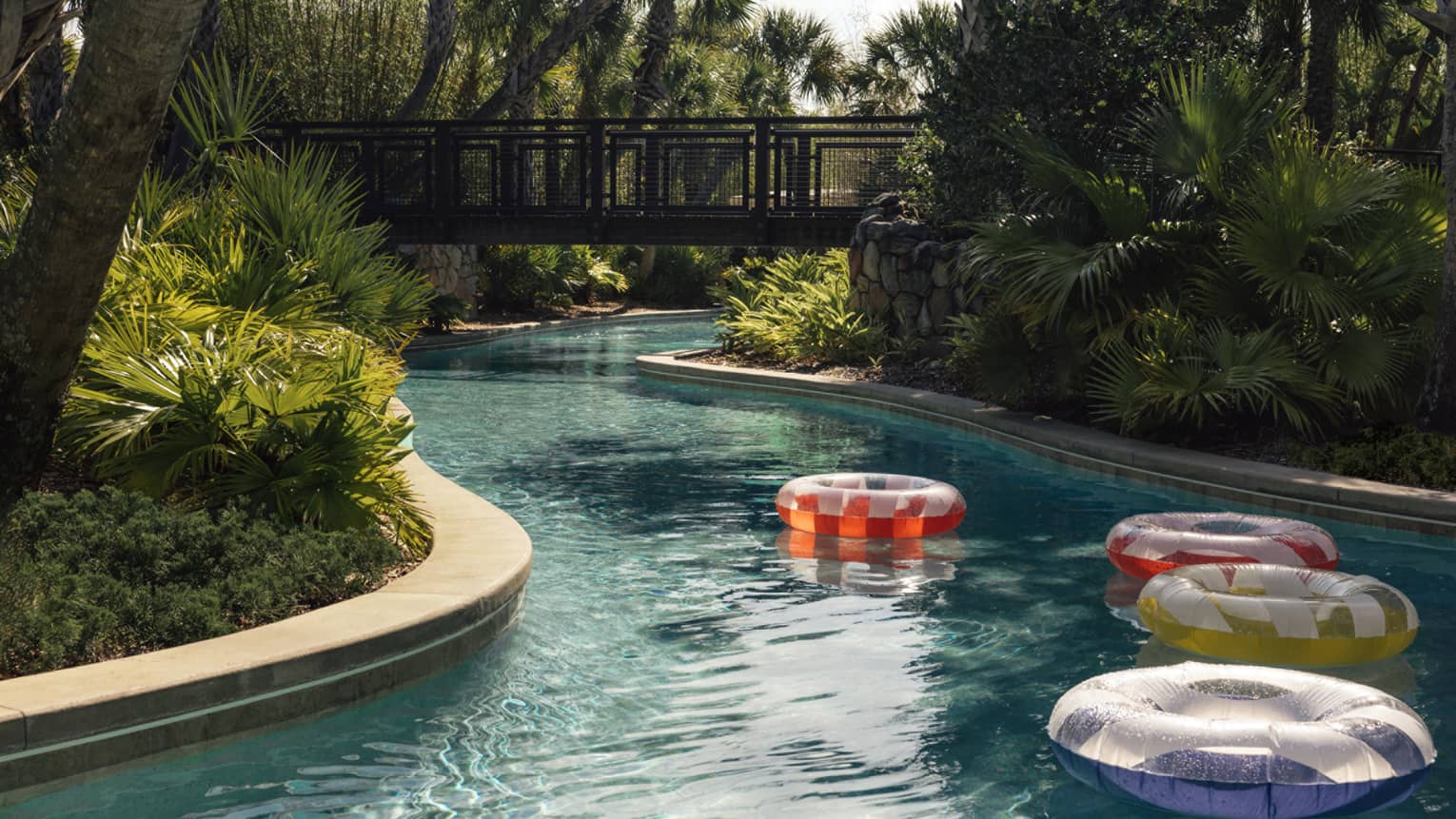Colourful inflatable pool tubes float on a winding pool bordered by lush trees. A walking bridge over the pool is in the background.