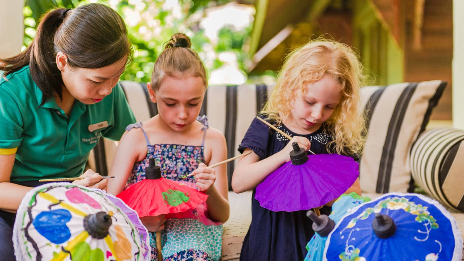 Kids Club staff helps young girls paint pictures on colourful paper umbrellas 