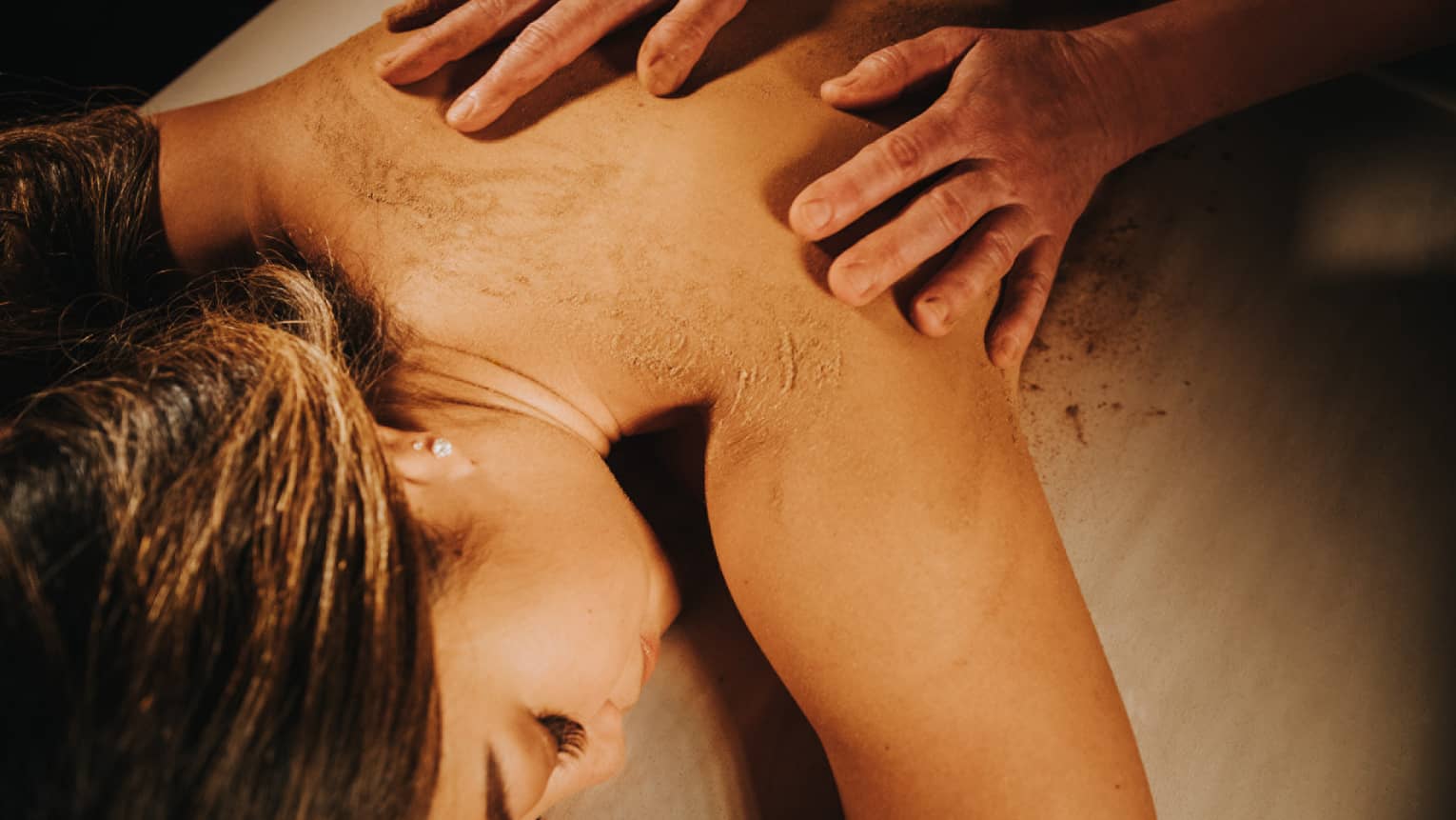 Two hands on woman's bare back as she lays on massage table in dimly lit room