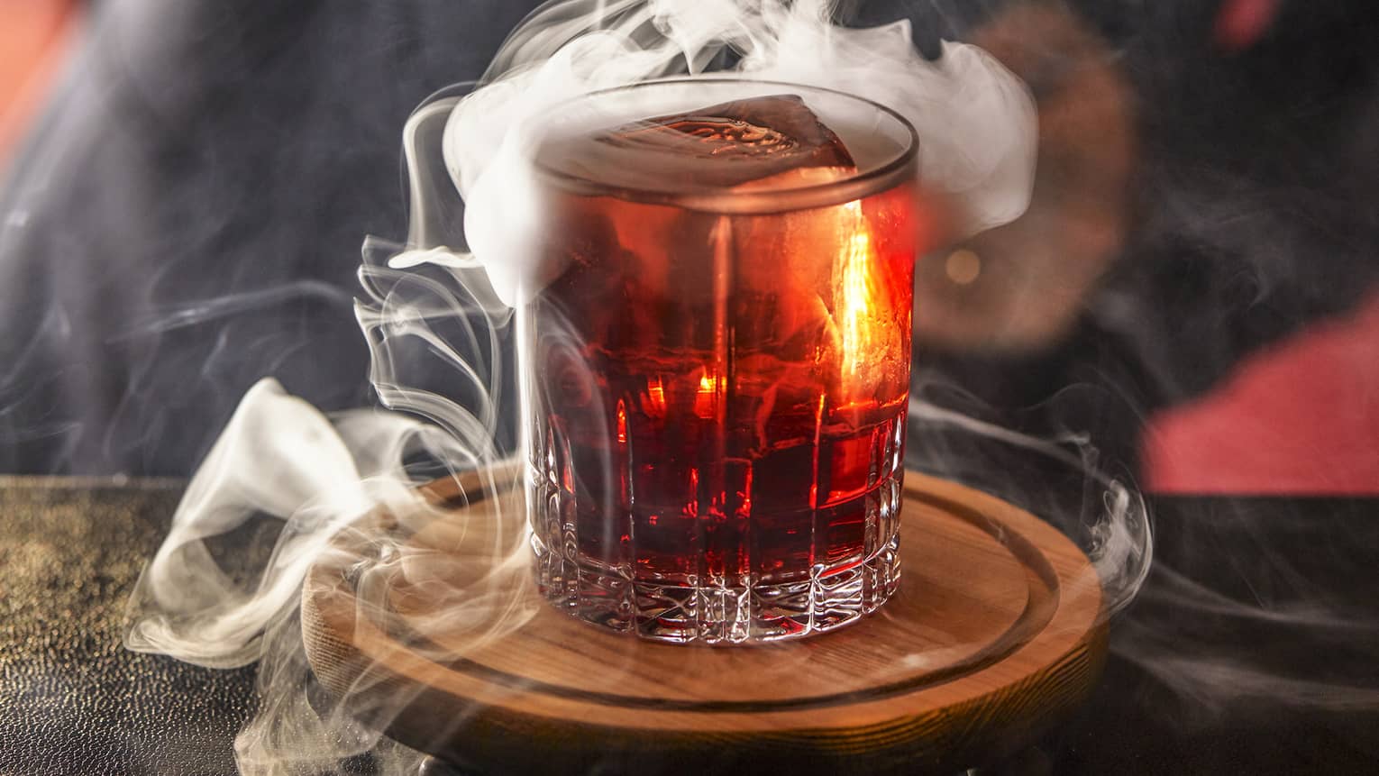 Bartender swirls smoke on top of a shot glass of amber-coloured liquor 