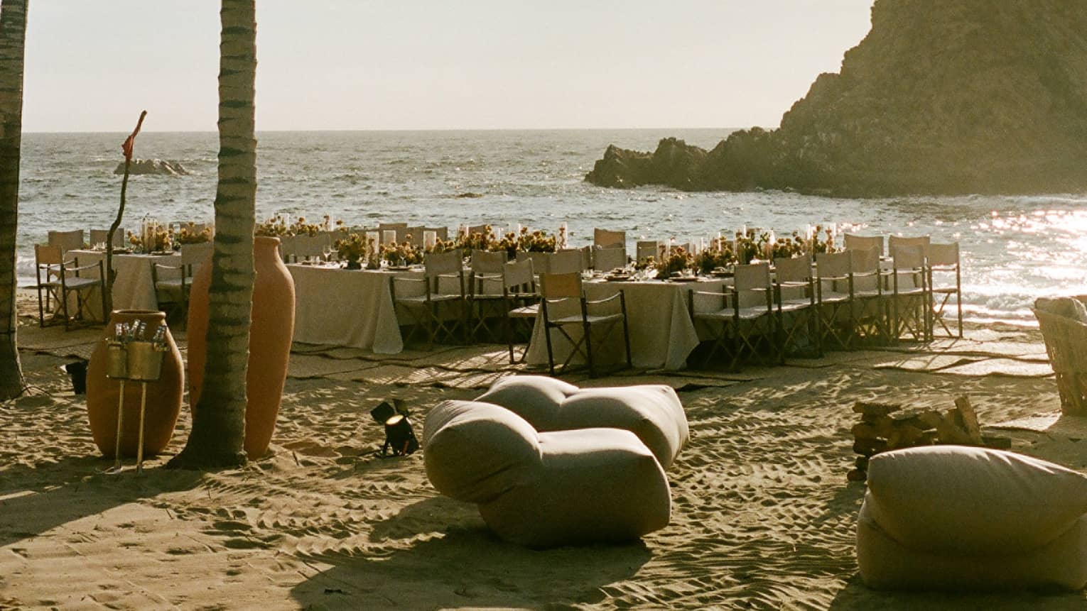 Dining tables for an event set near palm trees on the beach on a sun-drenched day, with a dramatic cliff in the background
