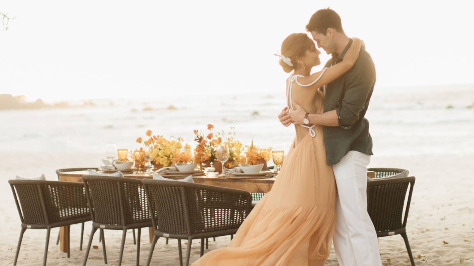 A man and woman hug next to a table on the beach decorate with orange flowers.