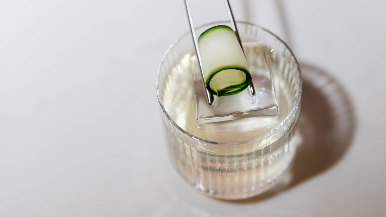 A thin, rolled up slice of cucumber is being placed atop a cocktail by a set of slim silver tongs
