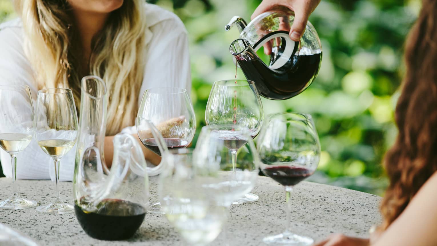 Smiling women at outdoor table, hotel staff pours red wine from artistic glass carafe