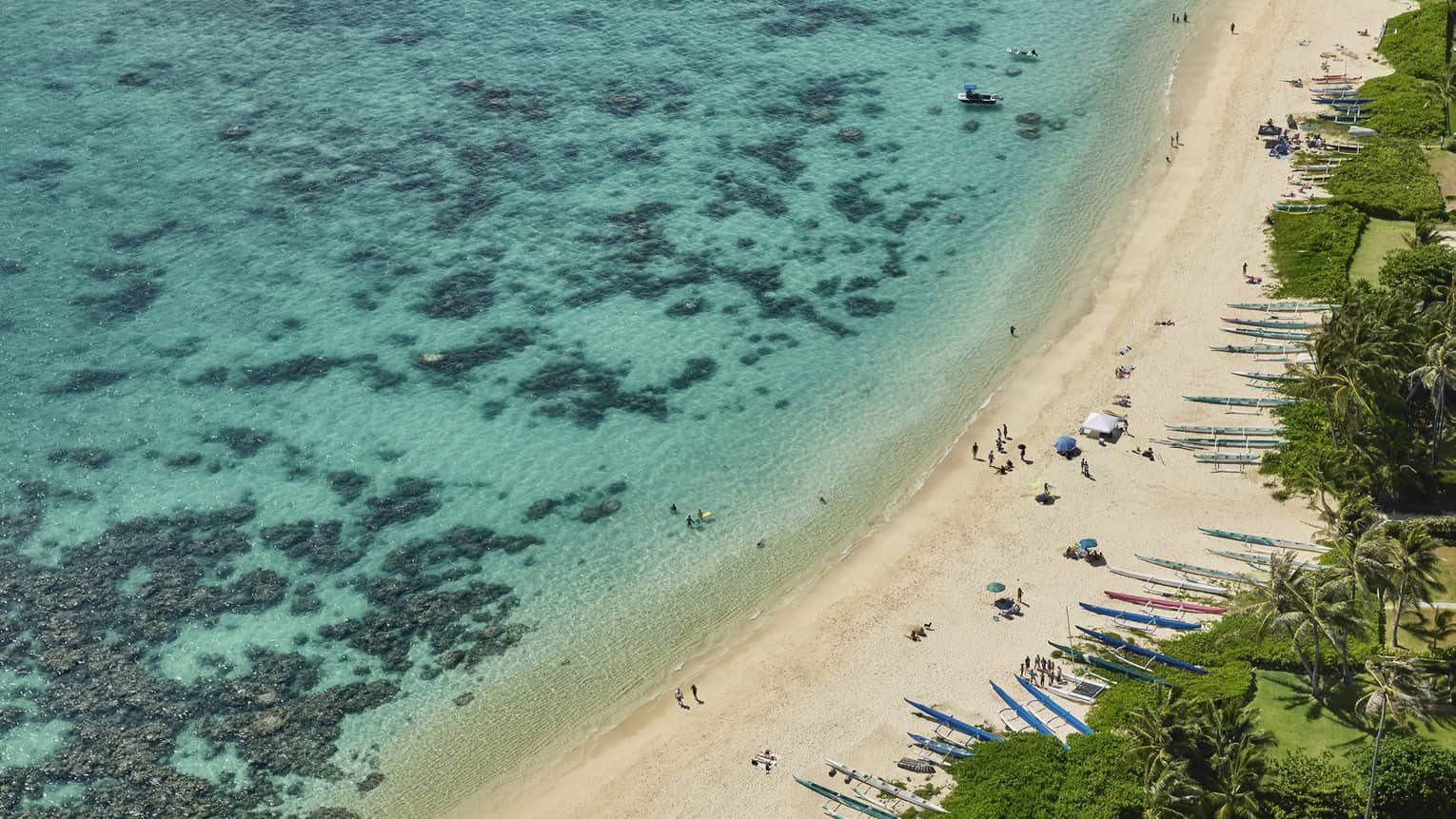 Aerial view of white sand beach lined with kayaks, blue lagoon with coral reef