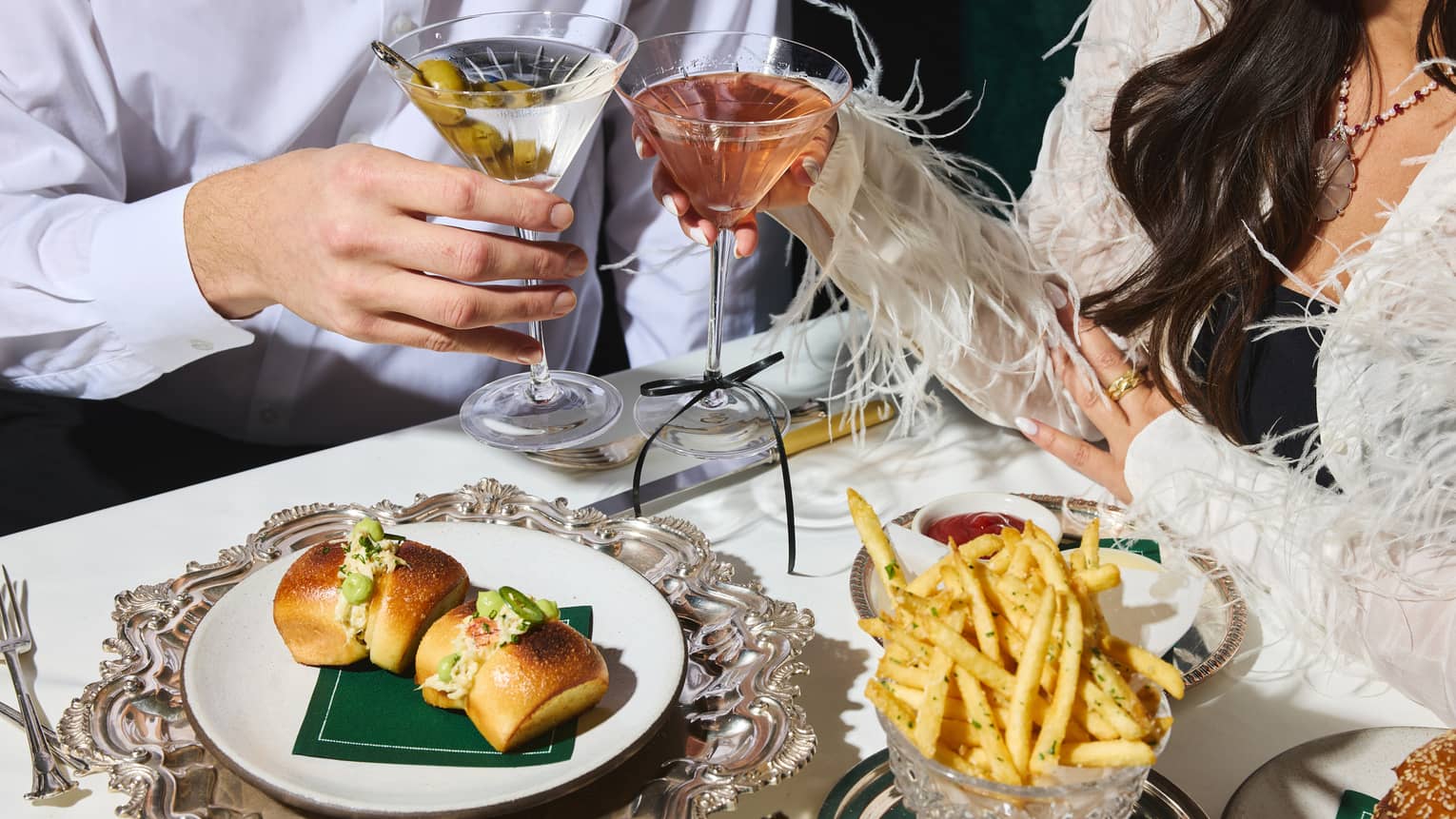 Two people toast their Martini glasses together over a dining table with French fries and small bites served on white plates ontop of silver platters