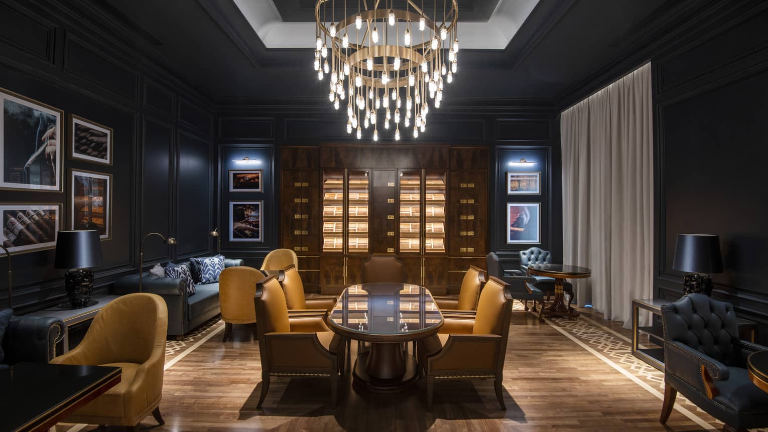 A lounge area with long wood table, dark sofas and walls and a hanging chandelier.
