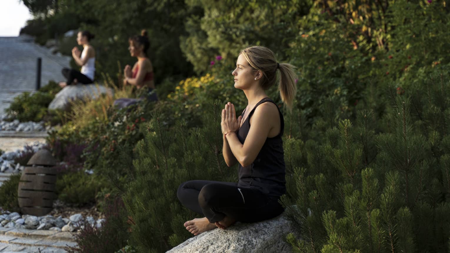 Three women outside in seated yoga pose on boulders separated by flowers