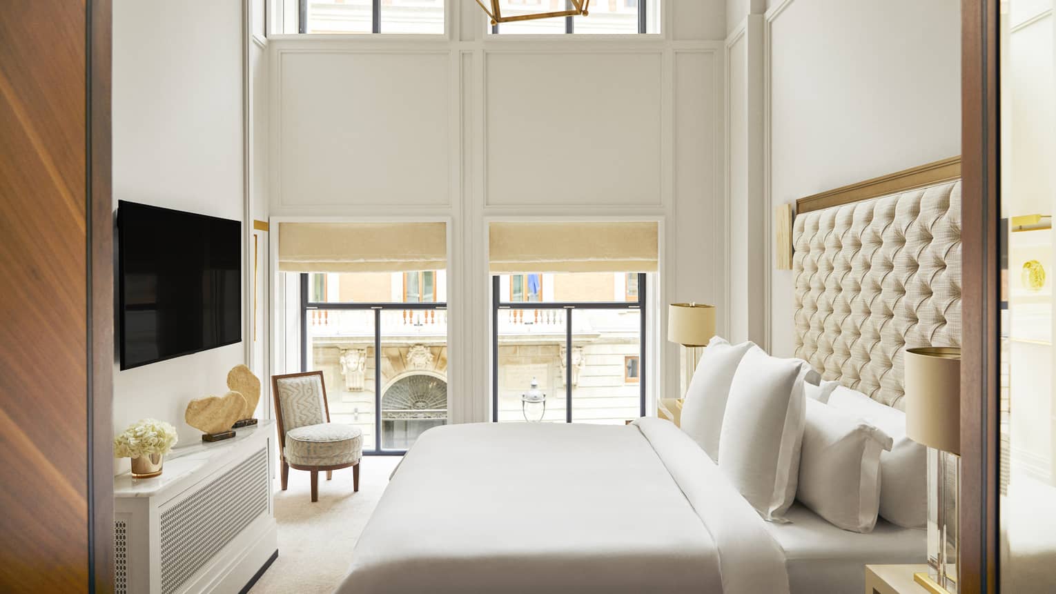 Hotel bedroom with a king bed with tufted headboard, wall-mounted TV, decorative console with stone sculptures, armchair and expansive windows with street views