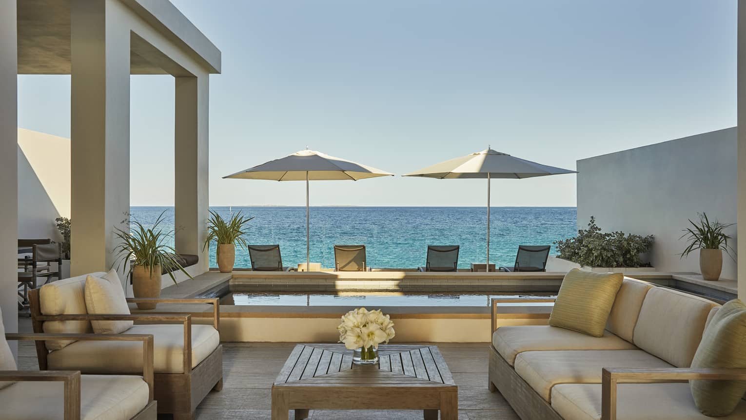 Outdoor patio living room with sofas, armchairs with white cushions, plunge pool, patio chairs and umbrellas, ocean