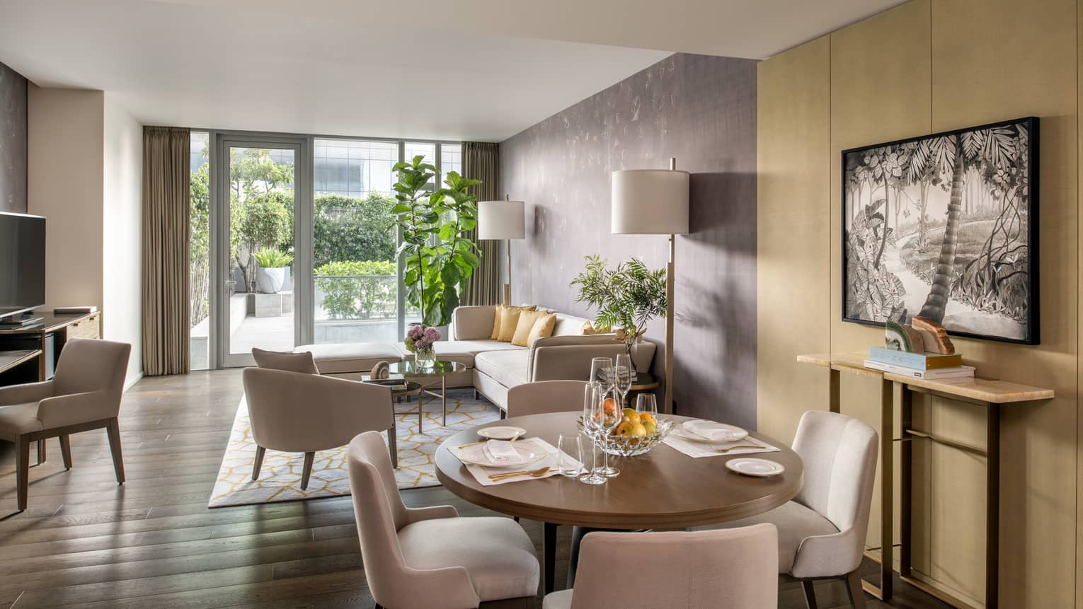 Guest room living and dining area with cream-coloured sectional sofa, round wooden dining table with four chairs, gold accents, floor-to-ceiling sliding glass door to patio