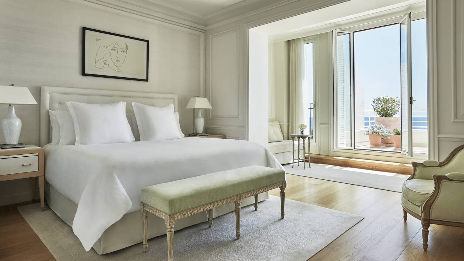 Hotel suite with white bed and green foot bench on neutral rug with terrace and sea views from tall glass terrace doors