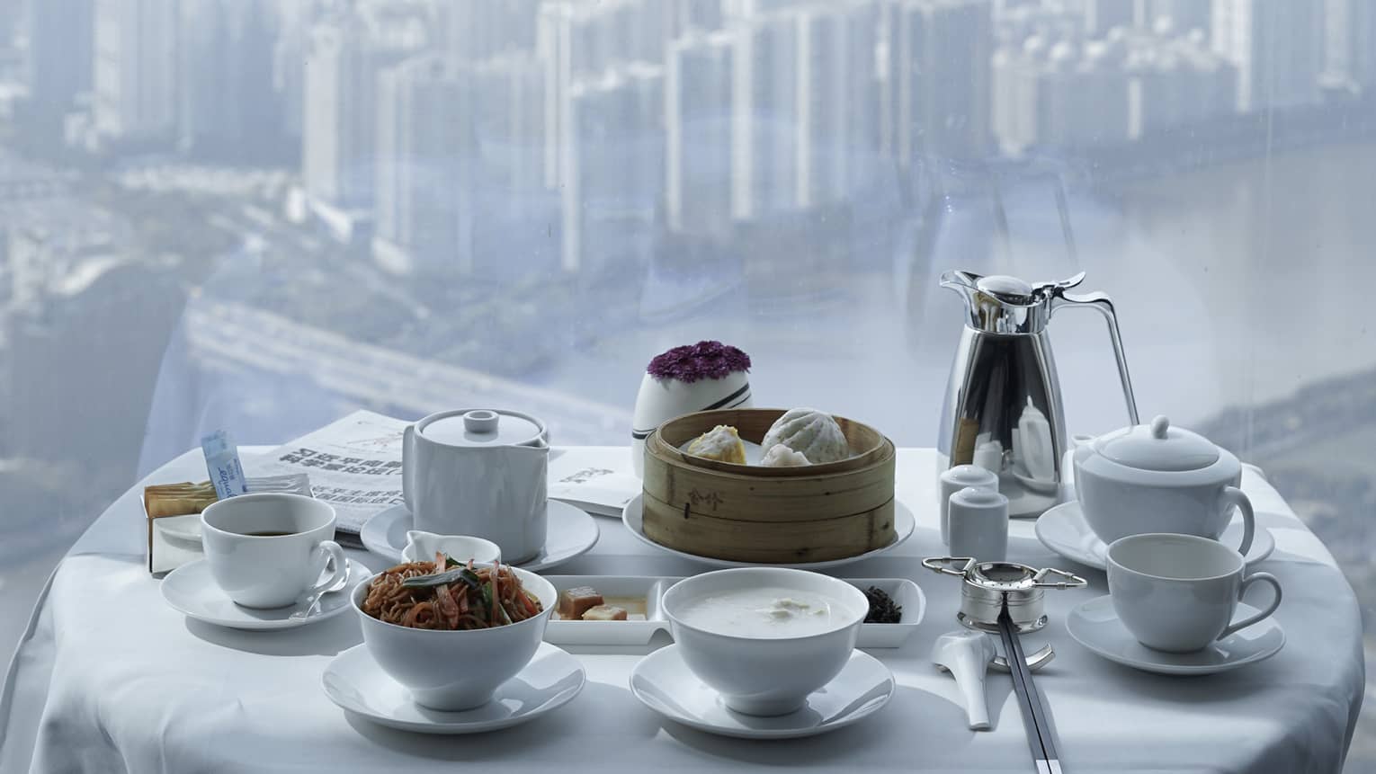 Small round table set with white dishes, crisp tablecloth and bamboo steamer. Dense cityscape under hazy sky visible below.
