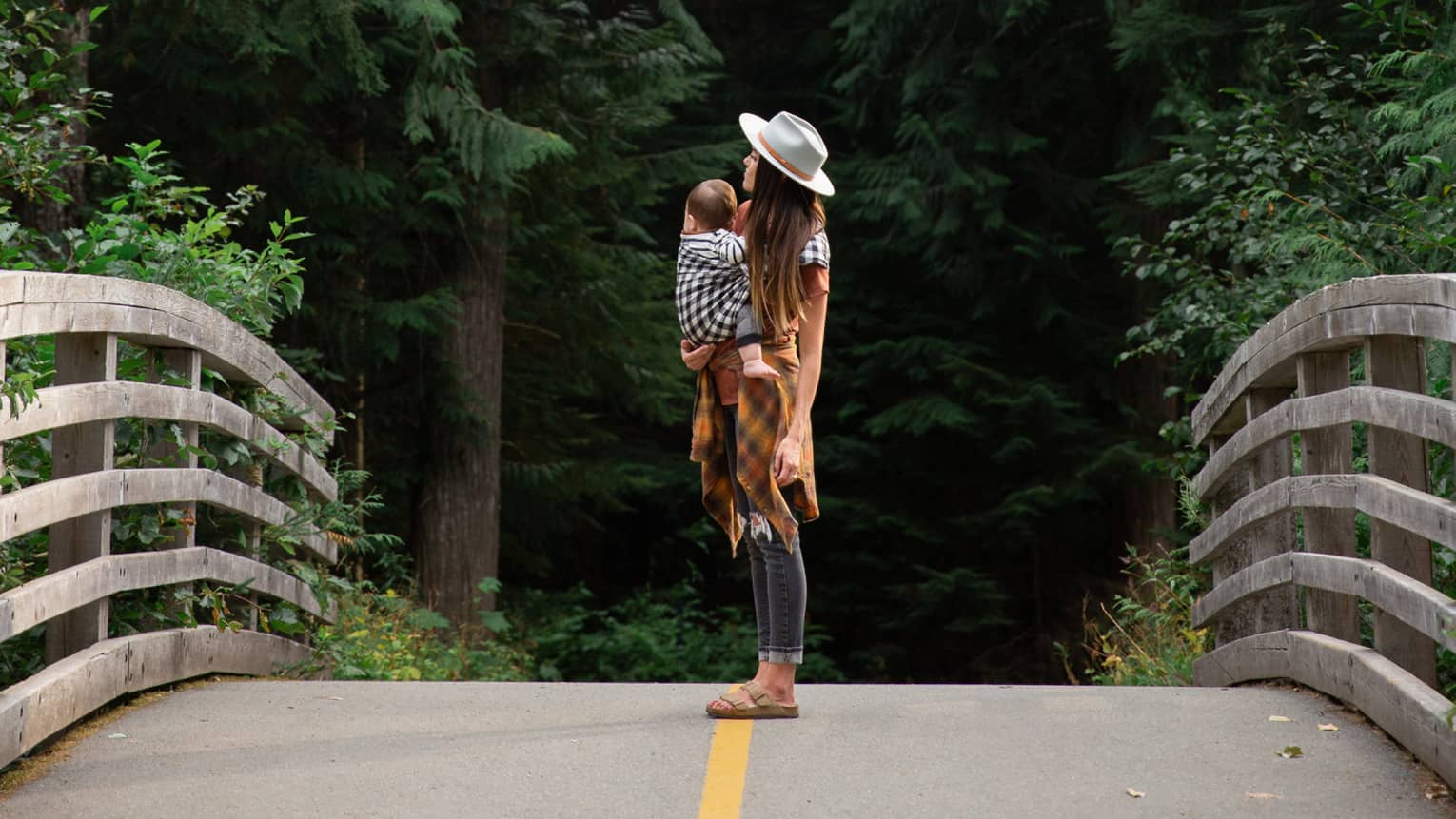 Woman with long dark hair wearing a beige hat, cognac-coloured long sweater and jeans holds a baby in a sling and stands in the middle of the bridge with tall green trees in the background
