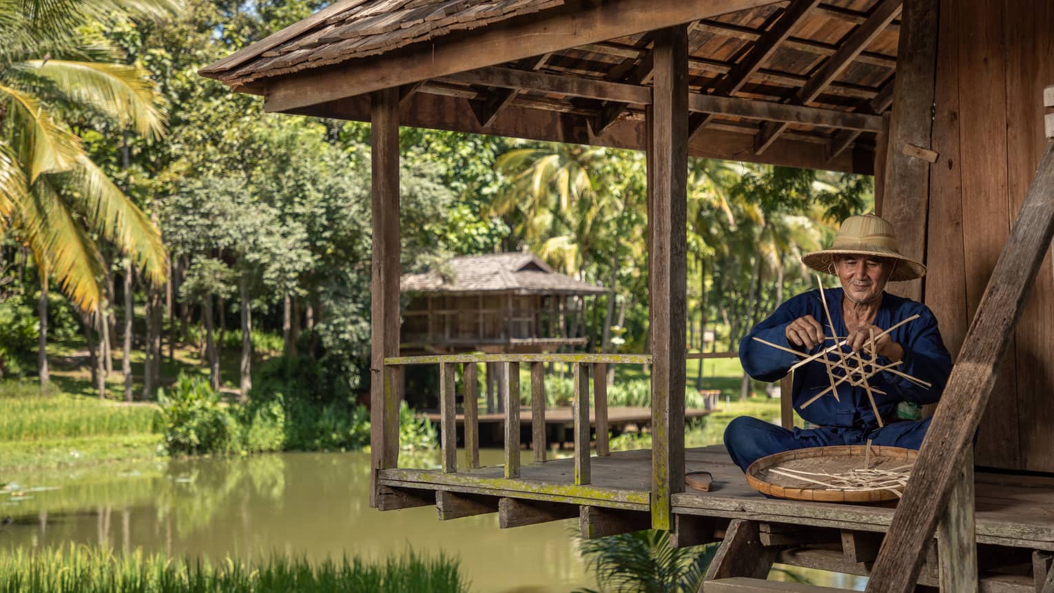 Man weaves a traditional Thai basket on the porch of a riverside pagoda