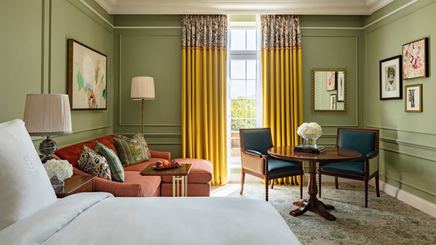 Mews Deluxe Room with King bed and white linens, red chaise lounge, accent table for two, avocado-coloured walls, floor-to-ceiling window with yellow drapes