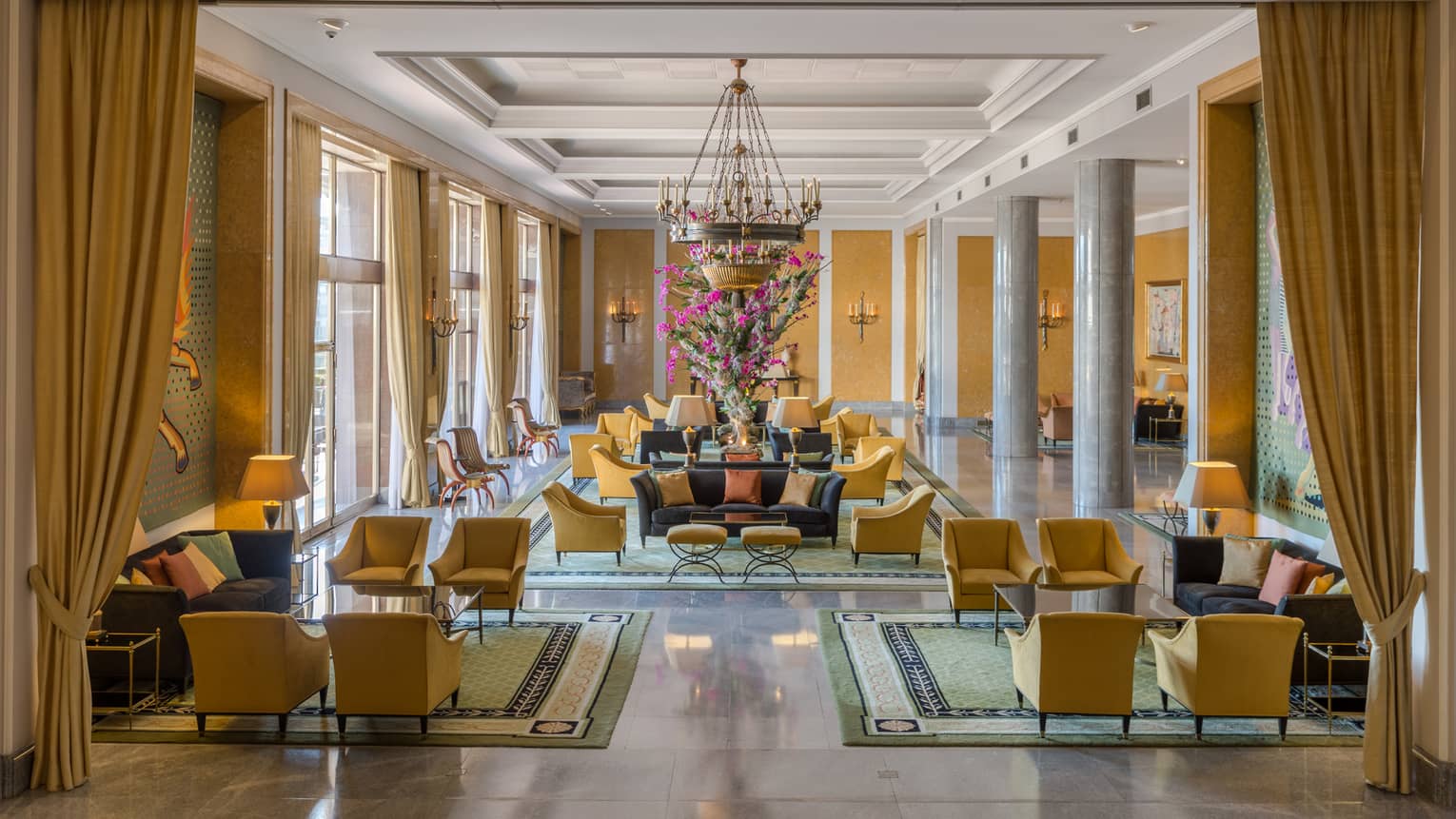 Bright hotel lobby with long gold curtains and armchairs, seating area under chandelier