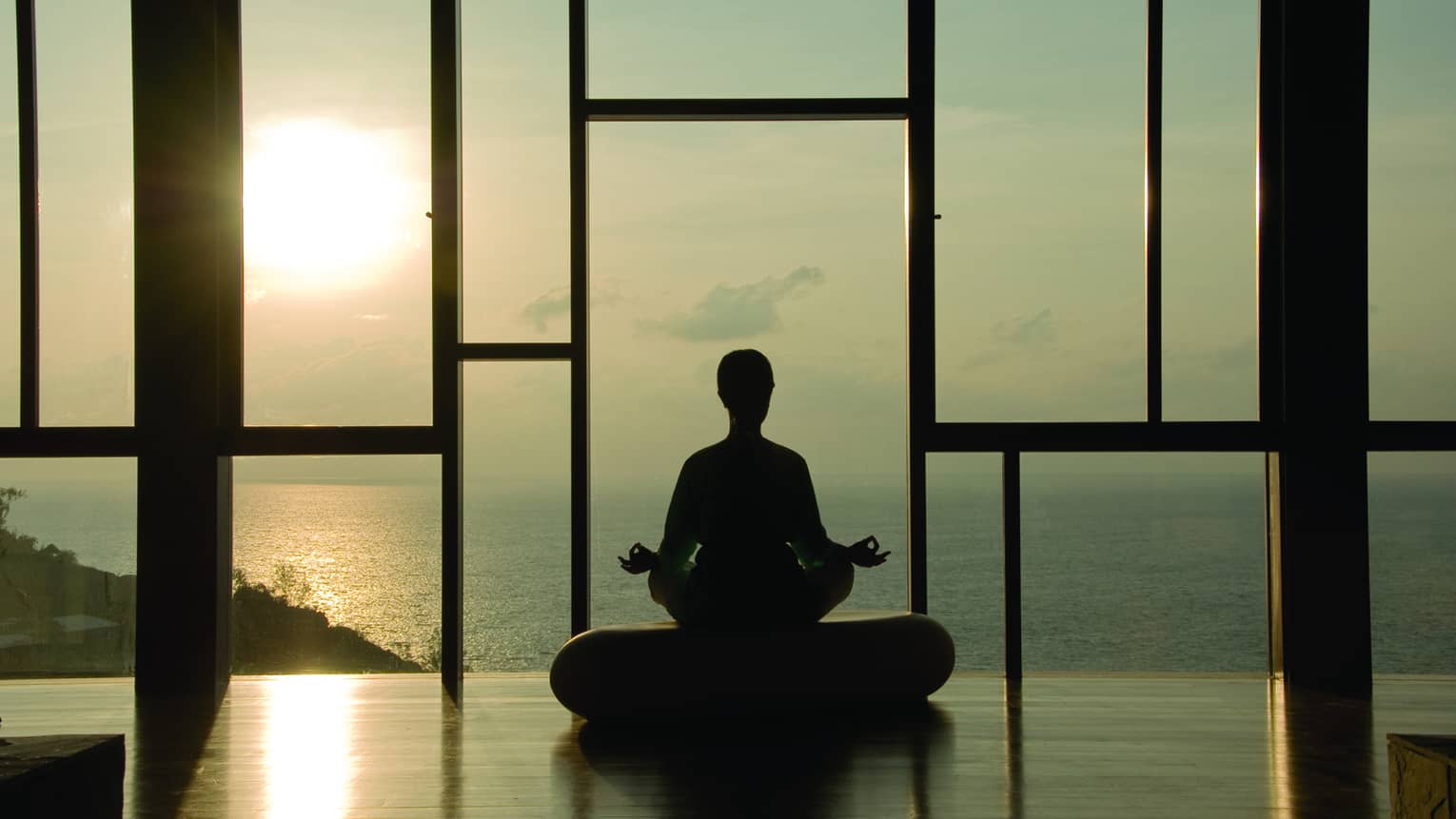 Silhouette of person meditating on cushion in front of floor-to-ceiling windows at sunset