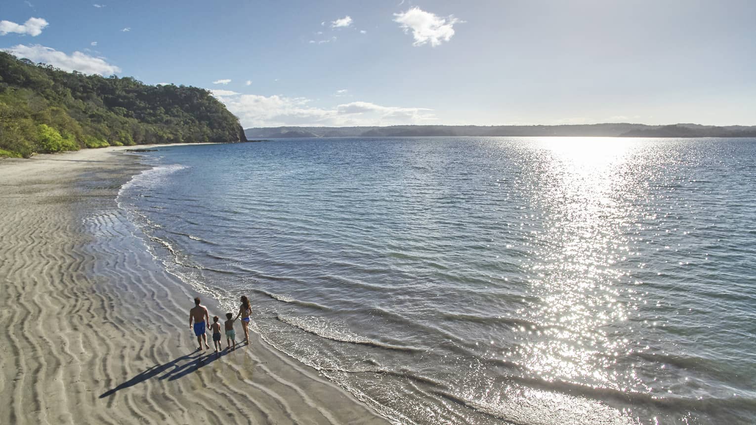 A family of four walks along the beach, under a high sun and a calm sea.