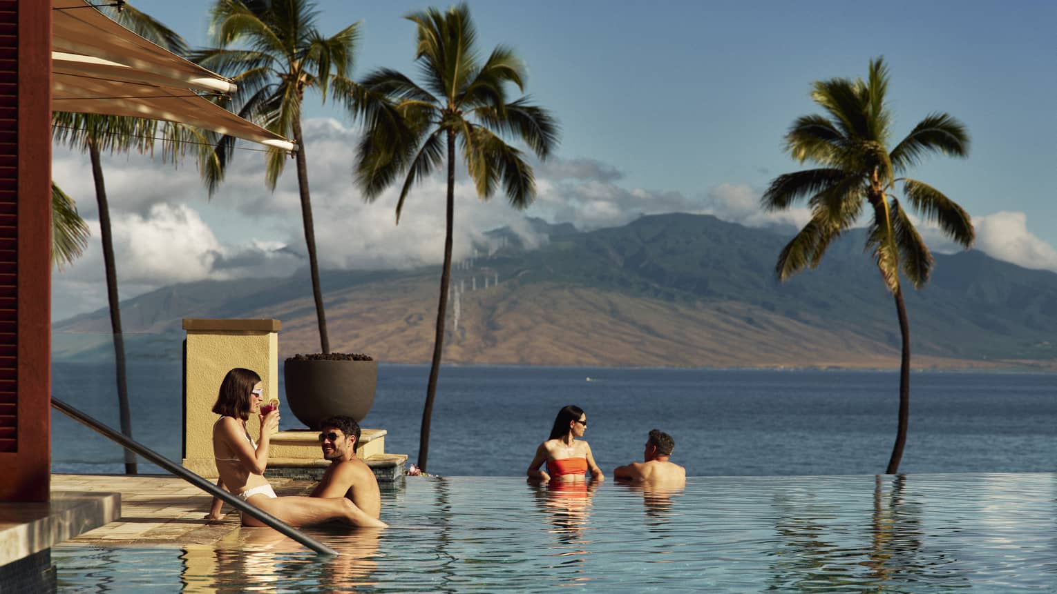 Four people relax in an infinity pool overlooking the ocean and palm trees with a mountain in the background