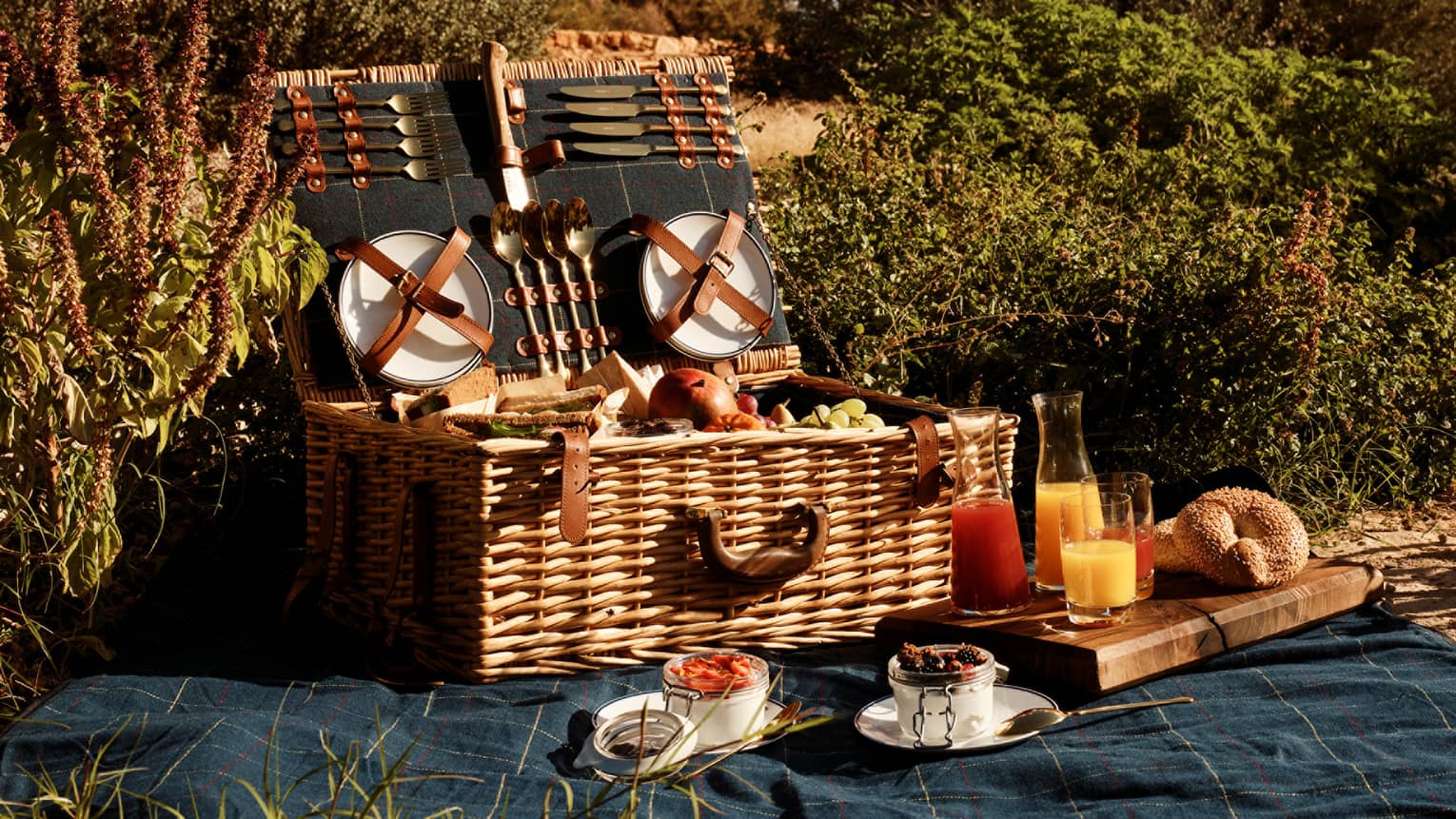 A large, well-stocked picnic basket with the lid open sits on a picnic blanket in an outdoor setting surrounded by greenery.