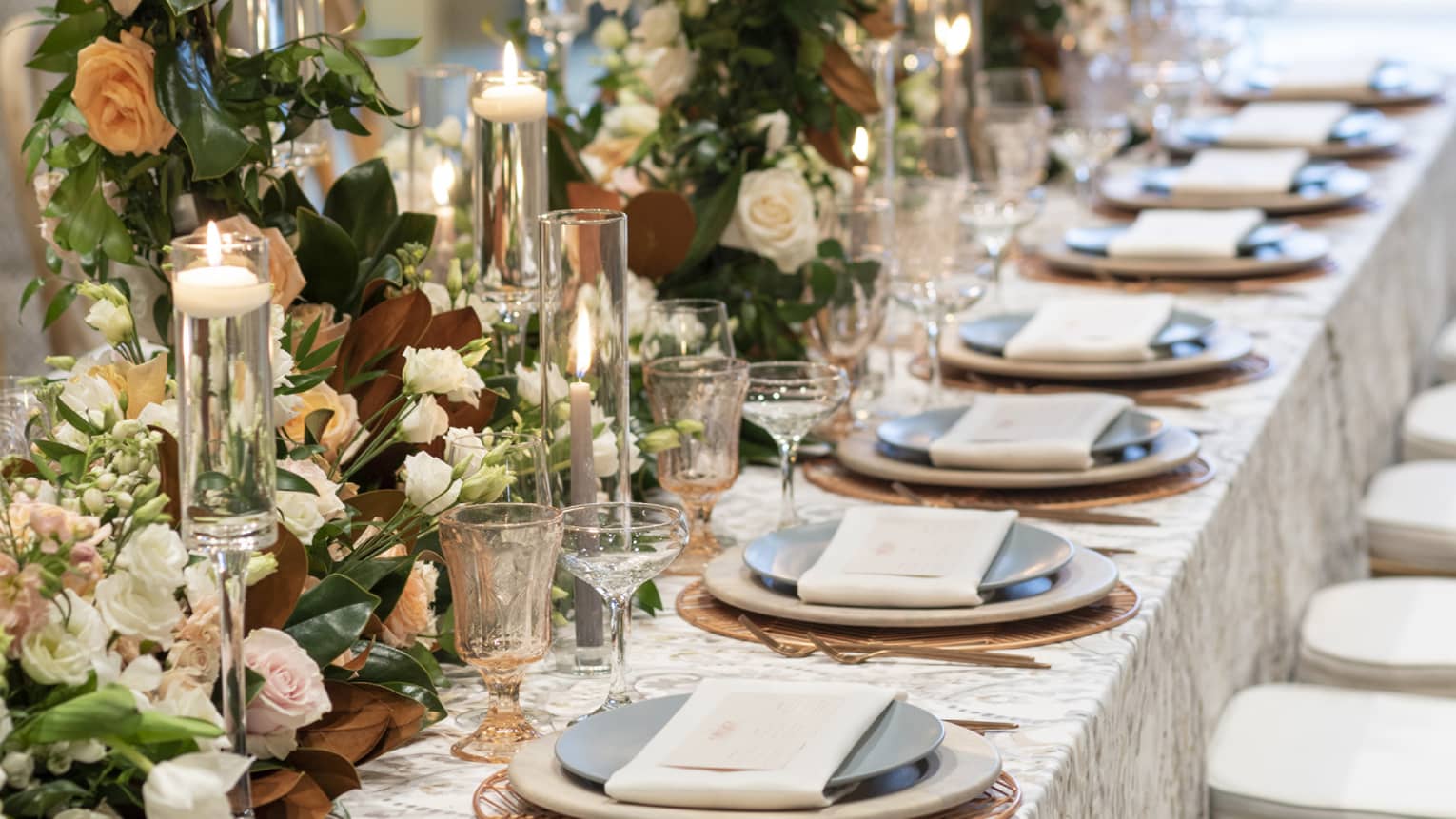An elegant wedding reception table set at Four Seasons Resort Orlando, bathed in natural light from large windows. The table is adorned with a luxurious floral runner featuring ivory blooms and lush greenery, complemented by glowing candles in tall glass holders. Fine china, crystal glassware, and crisp linen napkins await guests, promising a sophisticated dining experience.