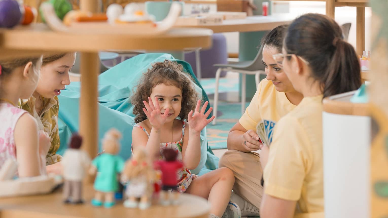 Three young children sit on the floor in a playroom playing cards with two adults