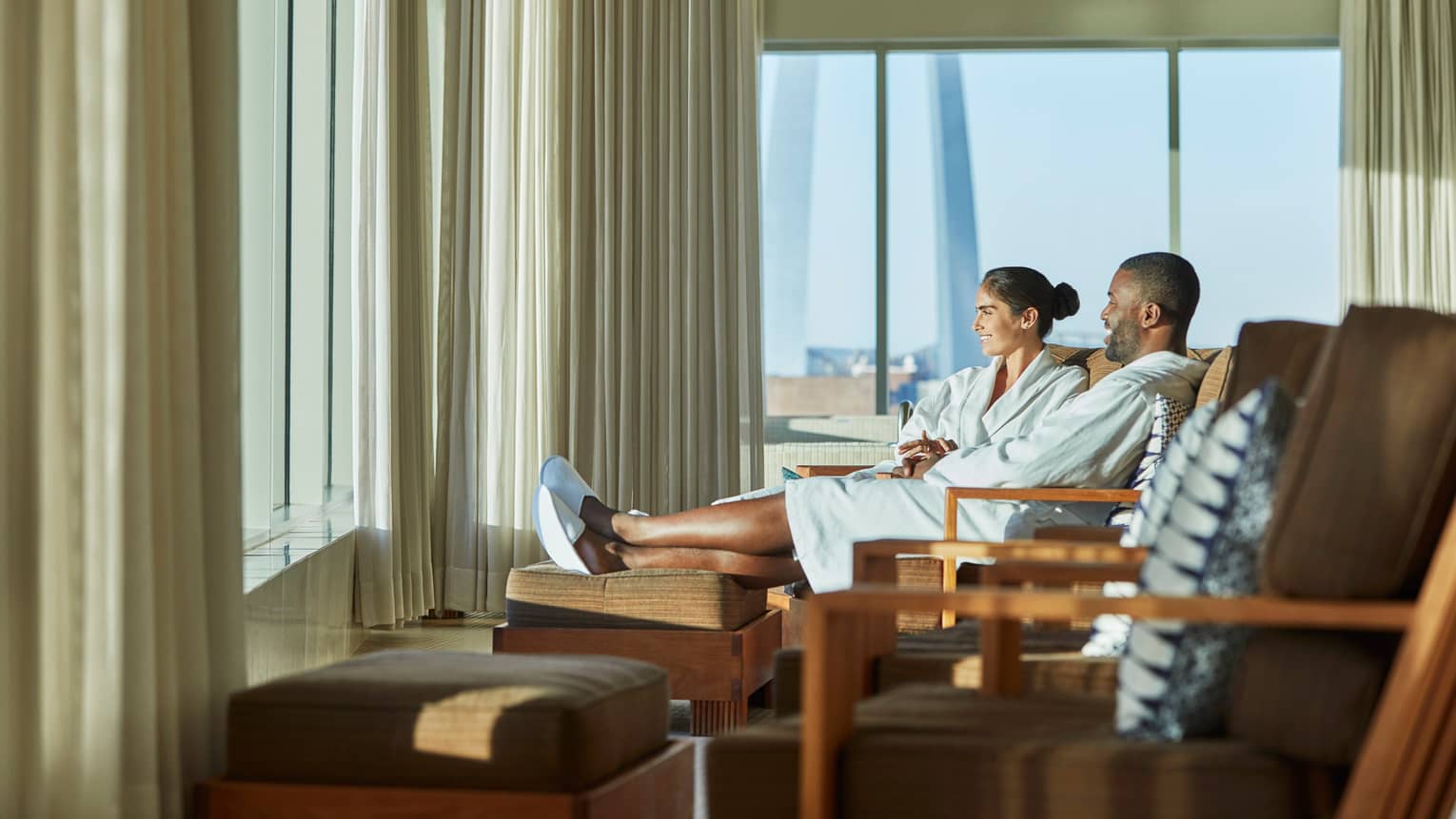 Smiling man and woman in bathrobes and slippers sit on lounge chairs near sunny window