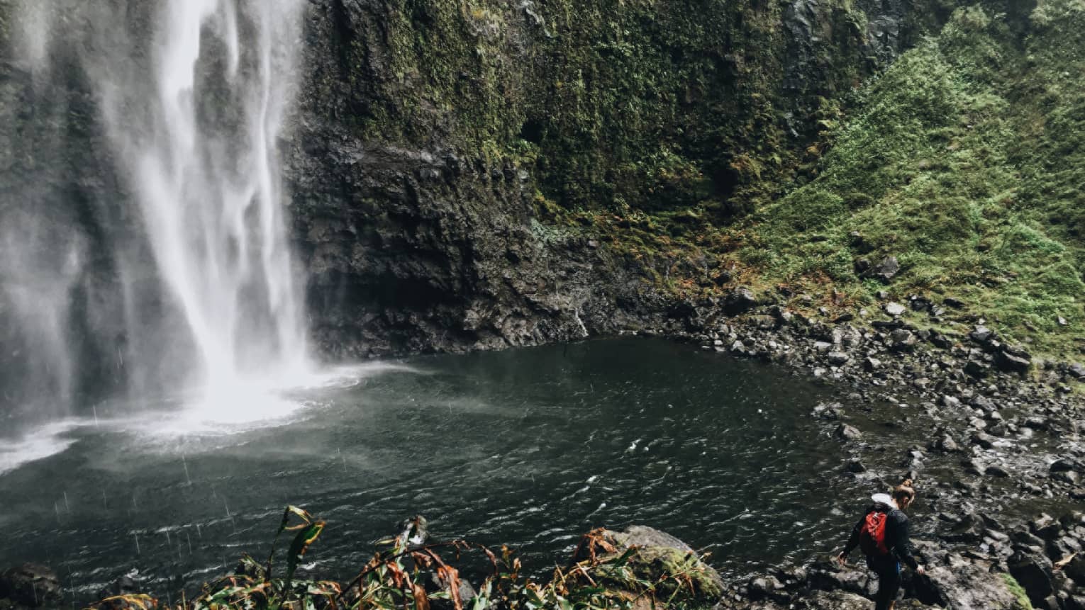 Back of woman hiking over black rocks under tall waterfall, small pond
