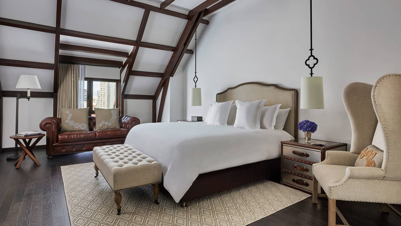 Brown leather loveseat in front of window, beamed ceilings sloped over bed, plush bench and armchair