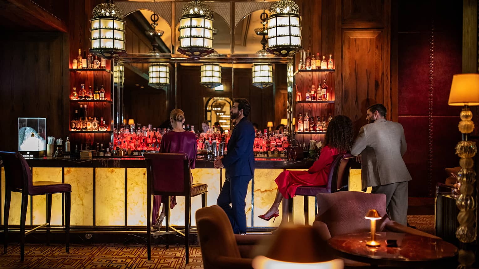 Two men and two women sit at an illuminated bar in front of several shelves of bottles.