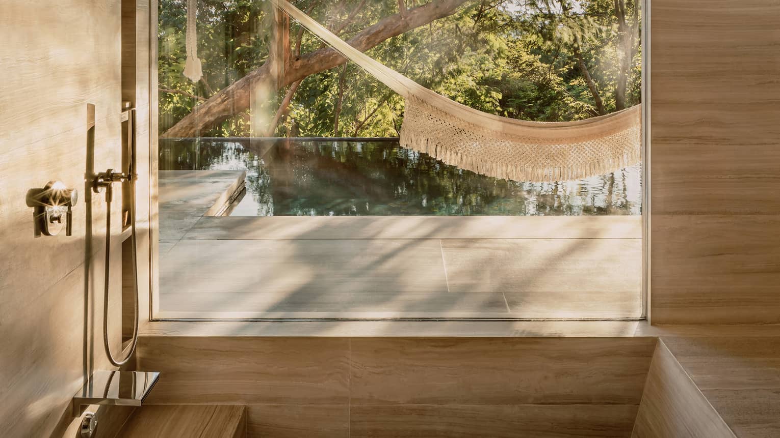 Minimalist bathroom with a deep tub and large window, offering a view of a hammock, infinity pool and lush trees outside, creating a serene, nature-inspired atmosphere