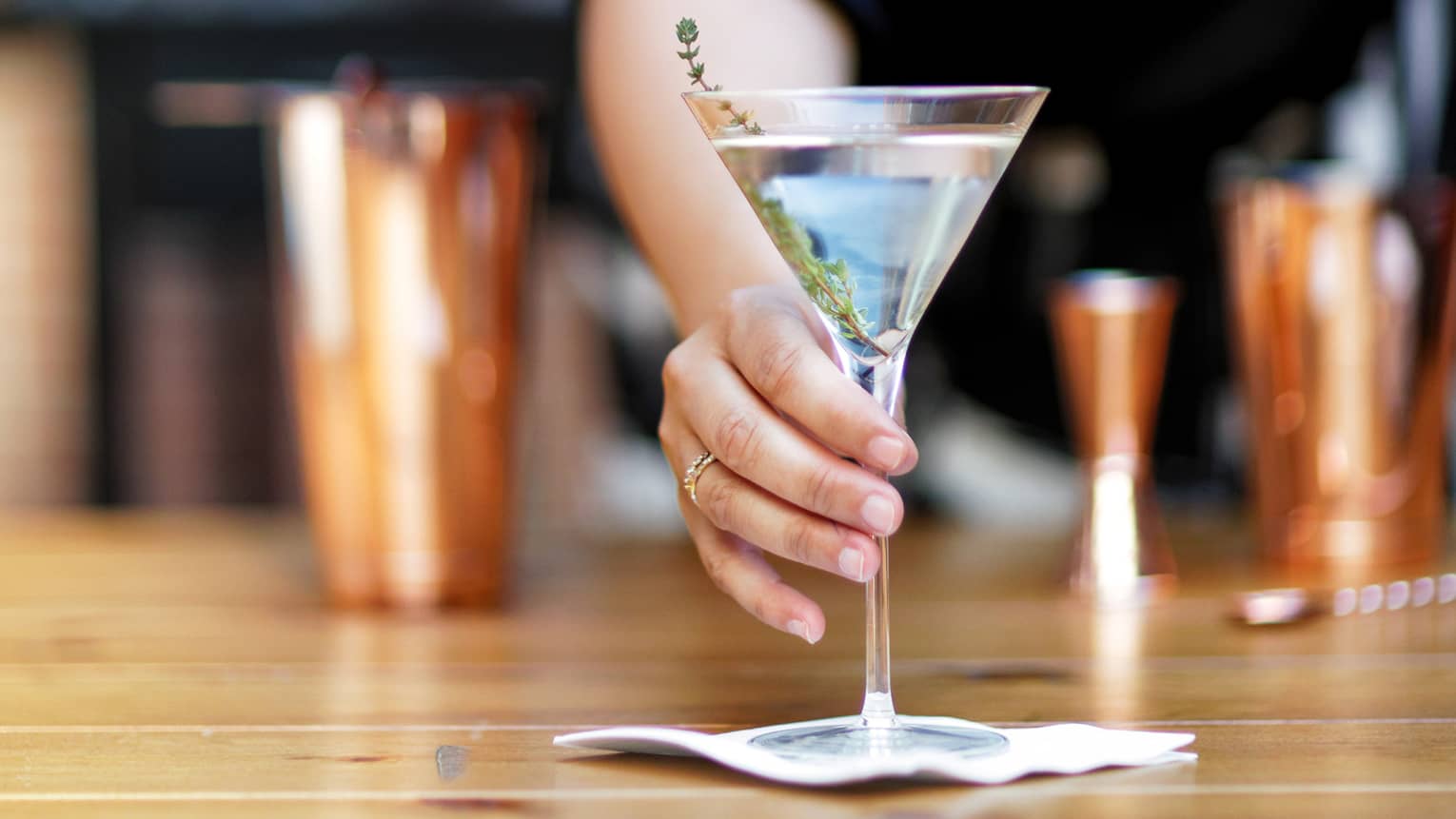 Bartender offering a martini garnished with a sprig of fresh thyme set on a small napkin, cocktail shakers in the background.