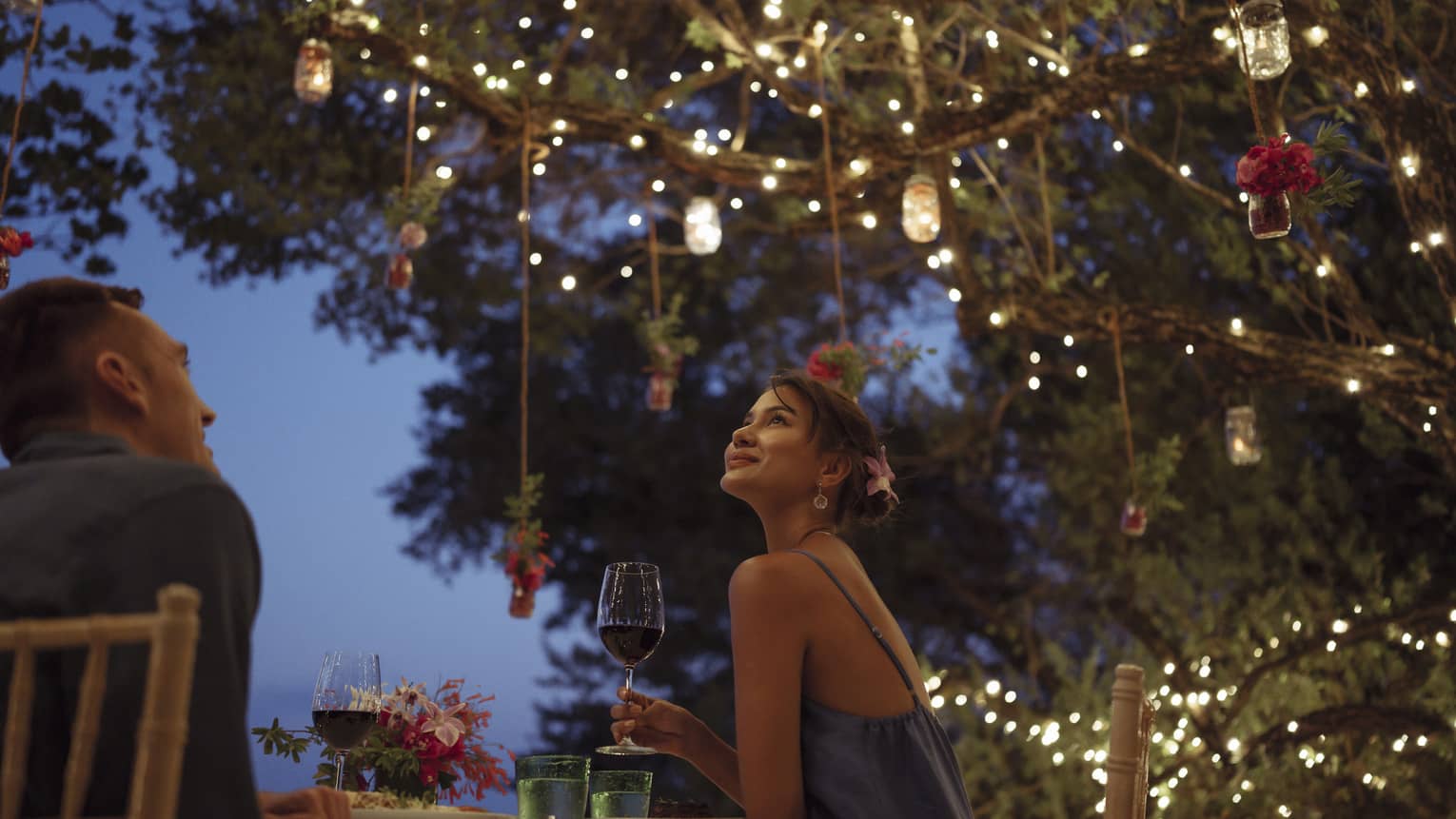 A smiling couple having a romantic dinner under trees covered in string lights and jars of flowers hanging from the branches.