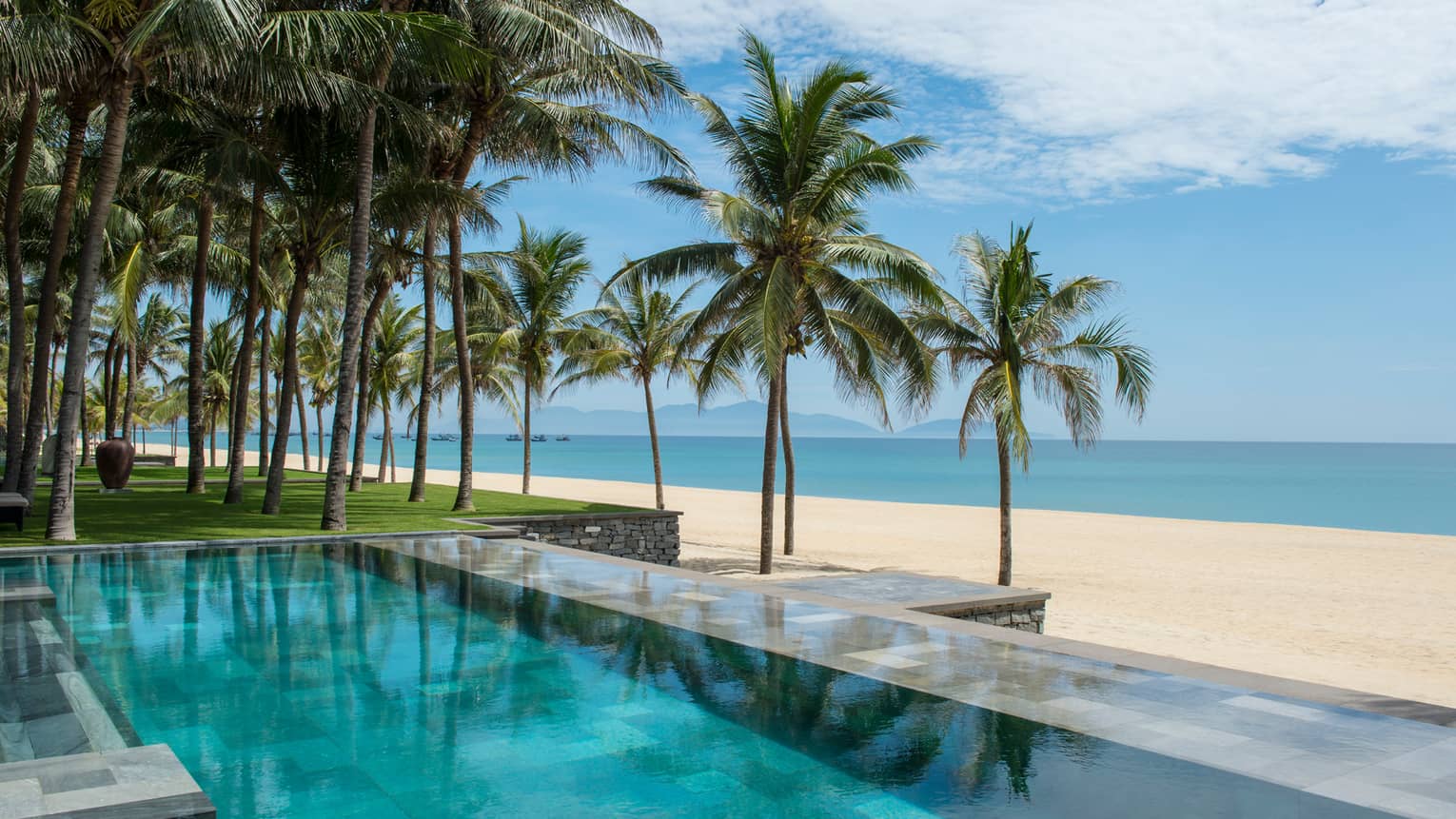Tall palm trees between stone outdoor swimming pool and white sand beach