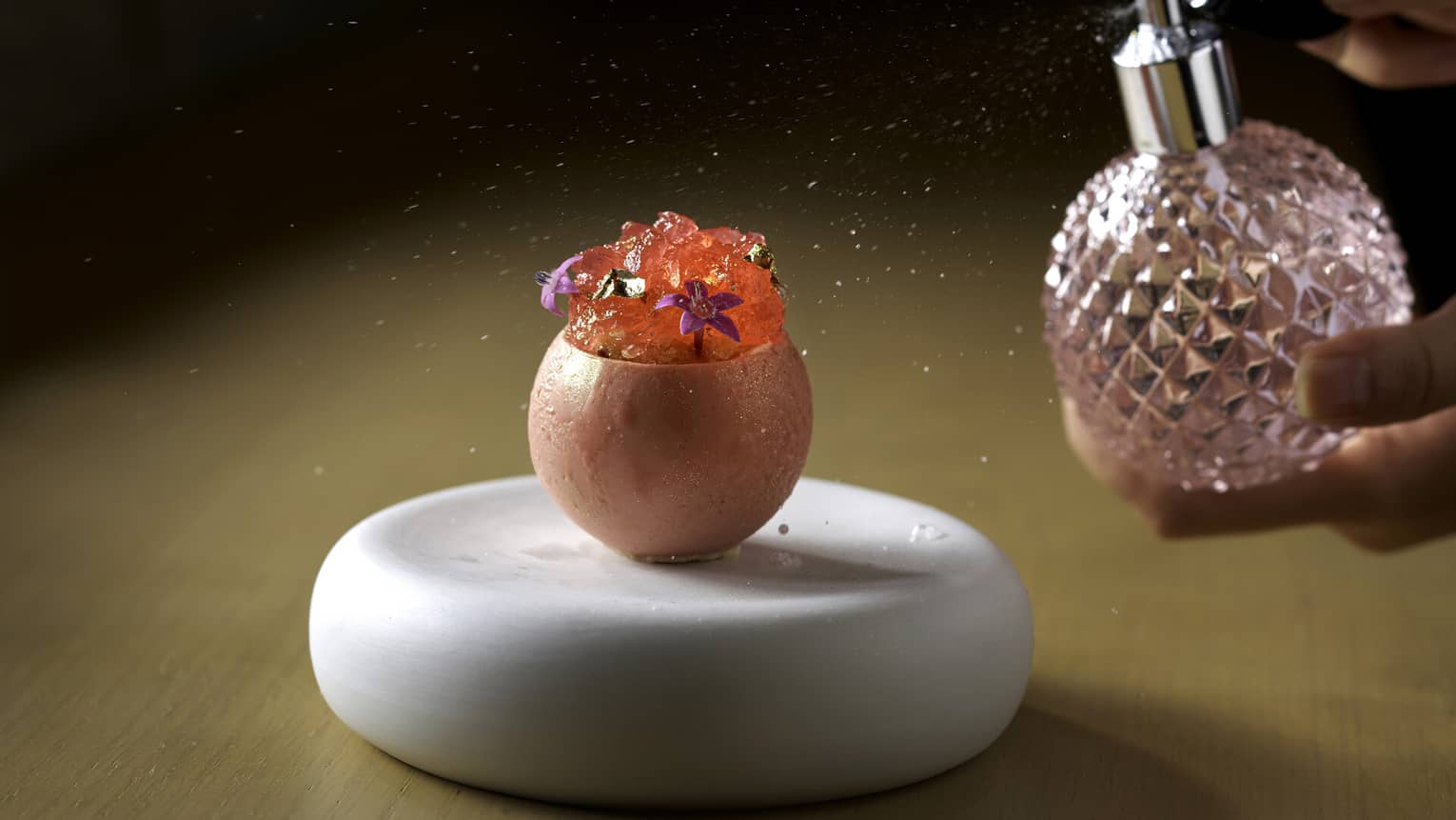 Person spritzing liquid from pink perfume bottle onto vessel filled with sake mousse