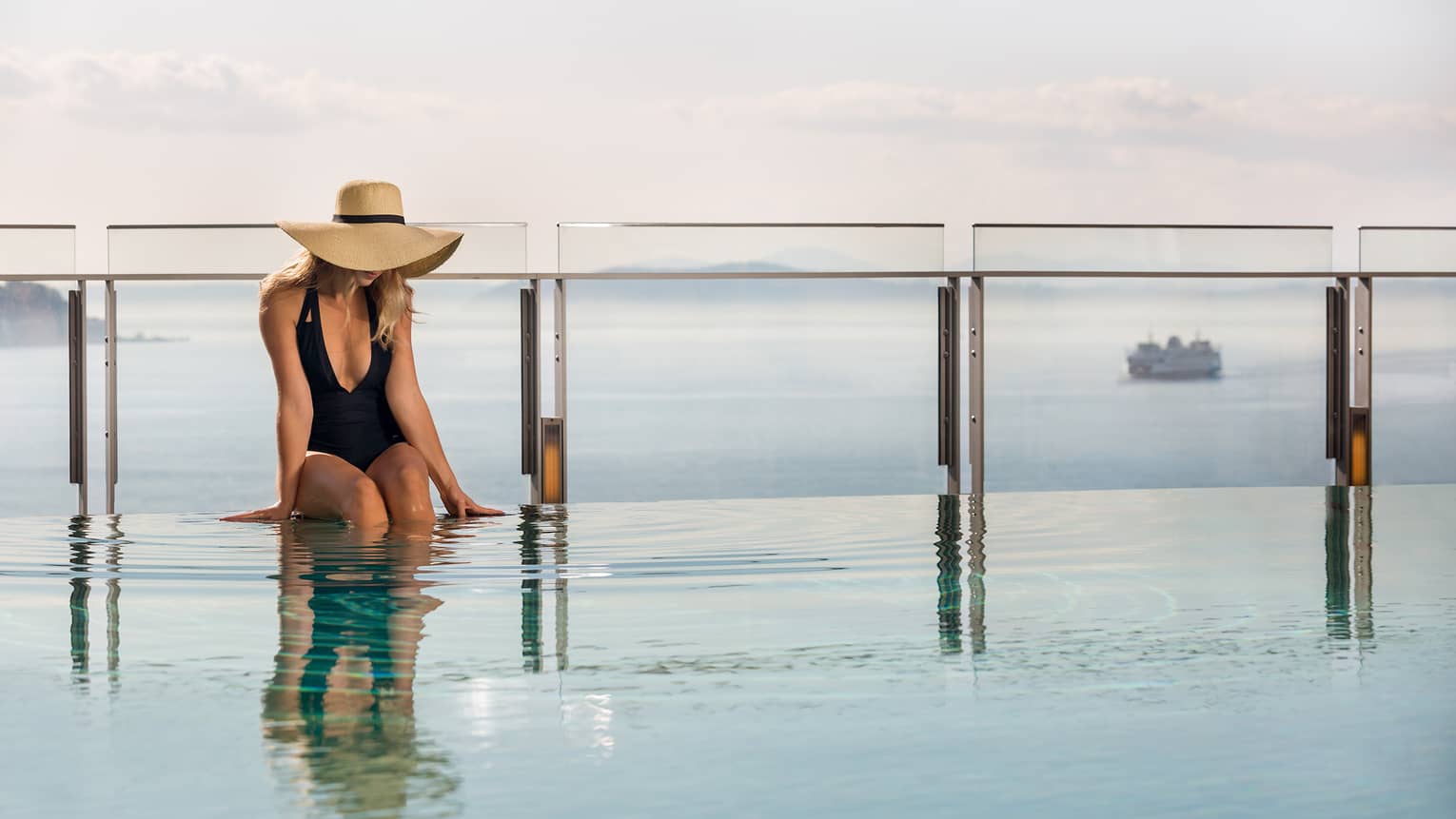 Woman wearing a black one-piece bathing suite and wide-brimmed hat, legs in the pool, sea in the distance