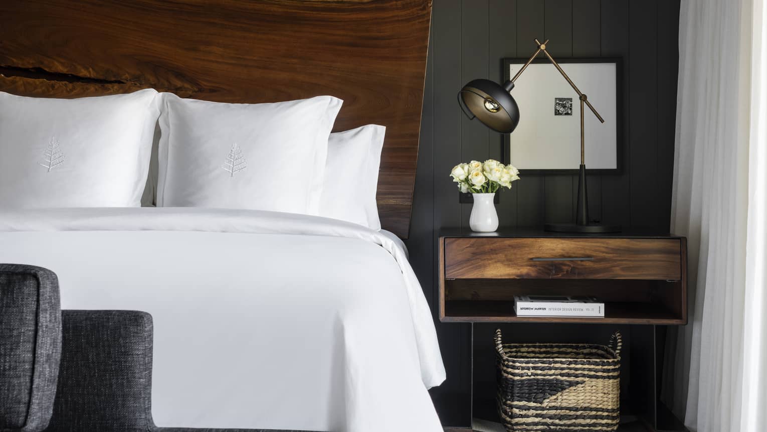 Close-up of a luxury resort white king bed with a dark wood headboard, wooden nightstand with a black lamp, vase of white roses and woven storage basket
