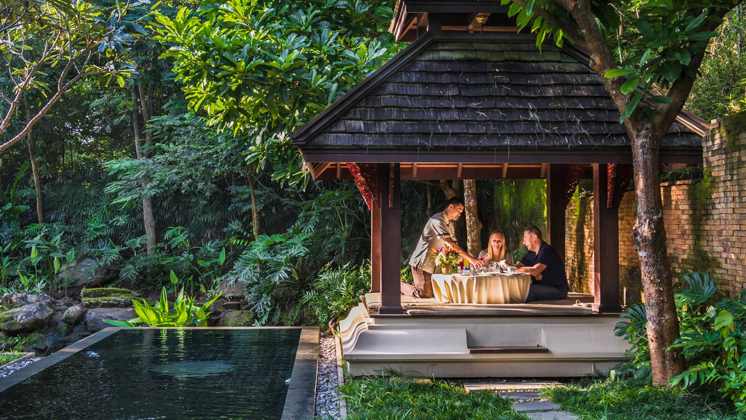 Hotel staff pours fresh juice for couple at private patio dining table under wood gazebo