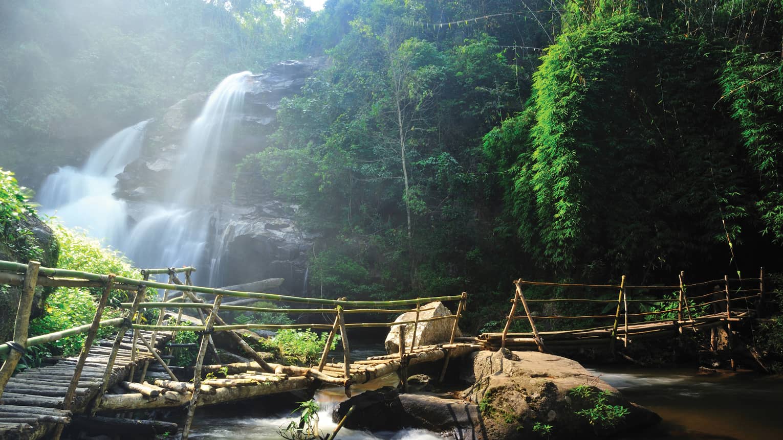 Rustic log bridge over river in front of cascading waterfalls at Queen Sirikit Botanic Garden