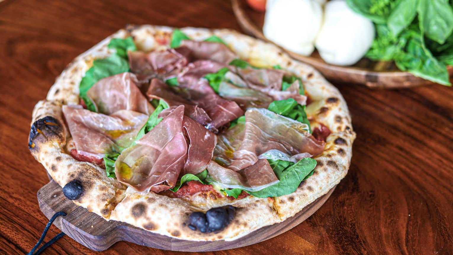 A flatbread on a wooden dish made with mozzarella cheese, tomato sauce, prosciutto di Parma, arugula and olive oil