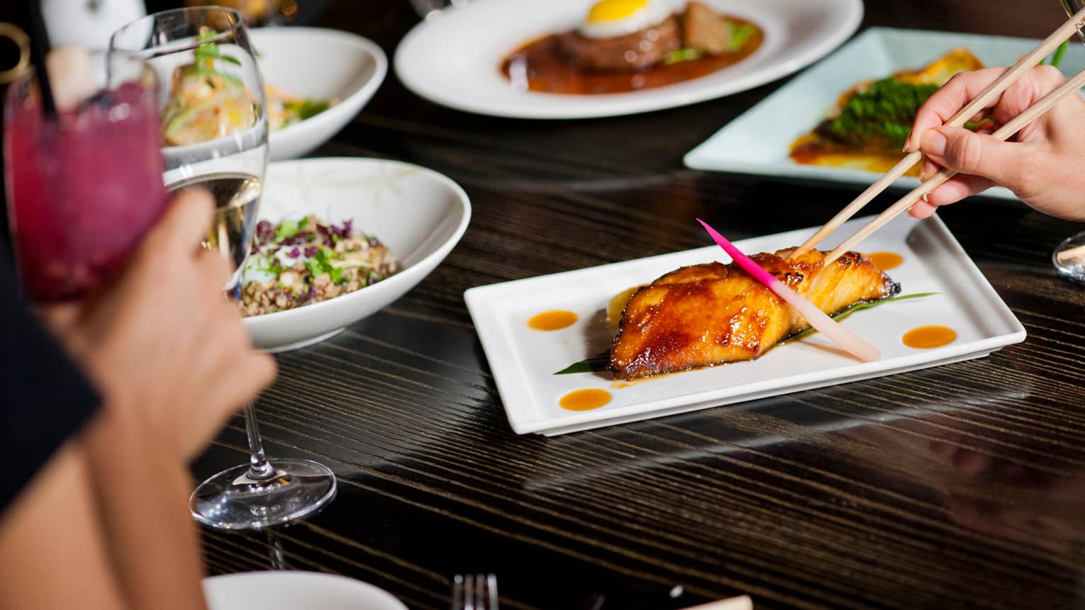 Close-up of hands, chopsticks by marinated grilled salmon on white dish on dining table