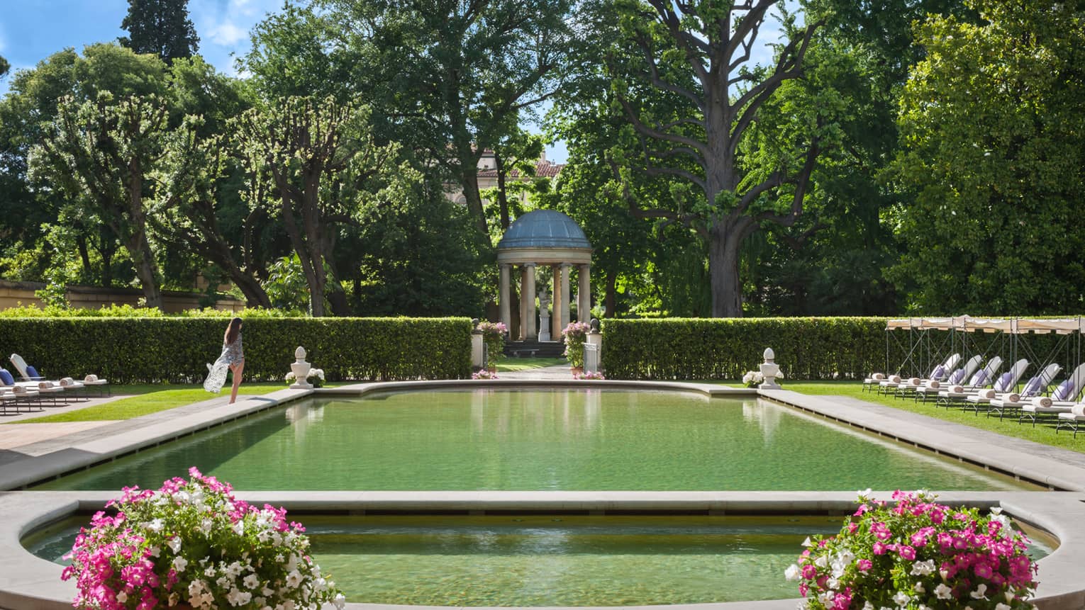 Woman in flowing robe walks past stone ledge of outdoor swimming pool surrounded by gardens