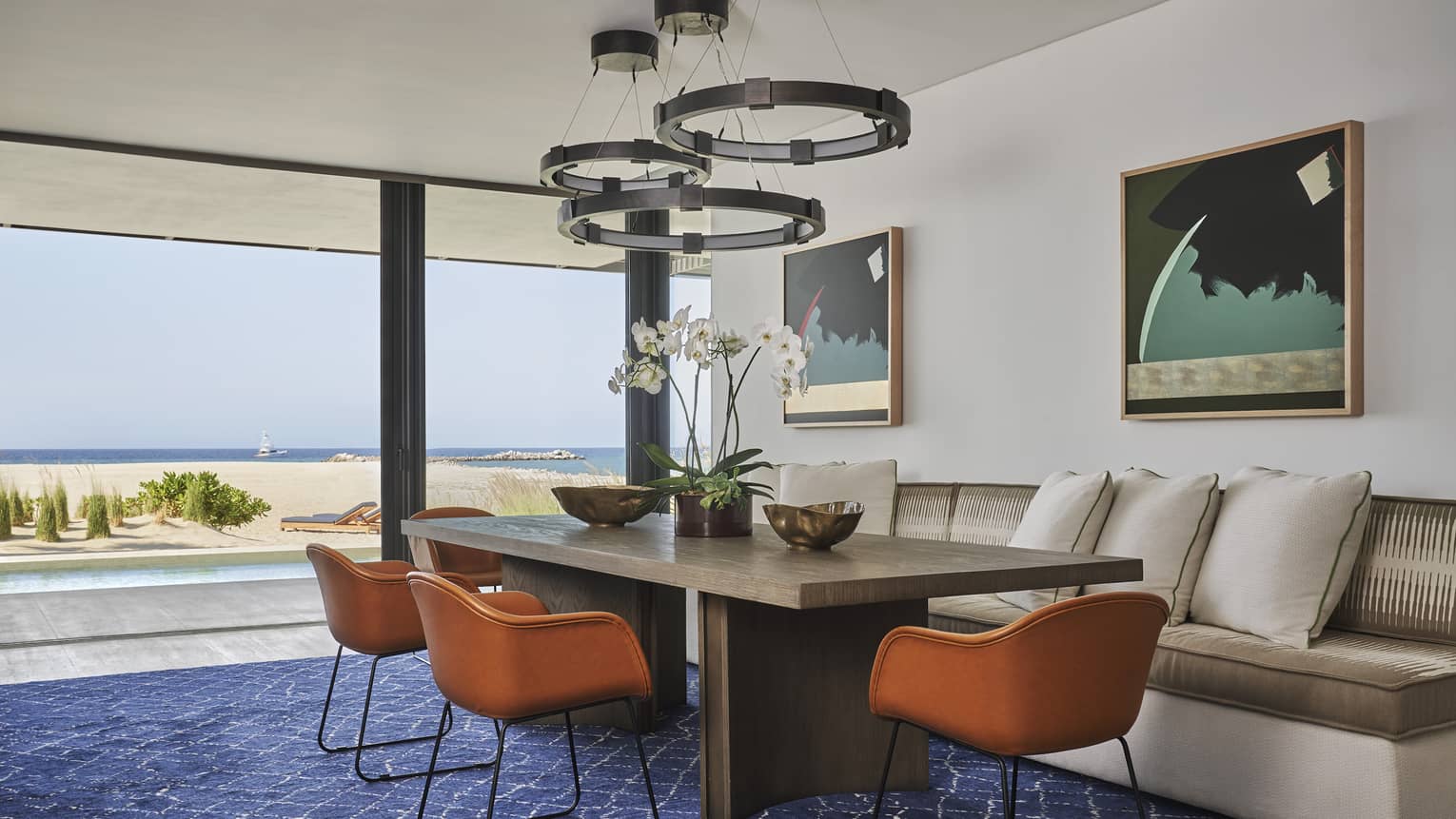 Two Bedroom Sunrise Suite dining area with rectangular table, wrought iron overhead lighting, cream sofa, floor-to-ceiling windows with ocean views
